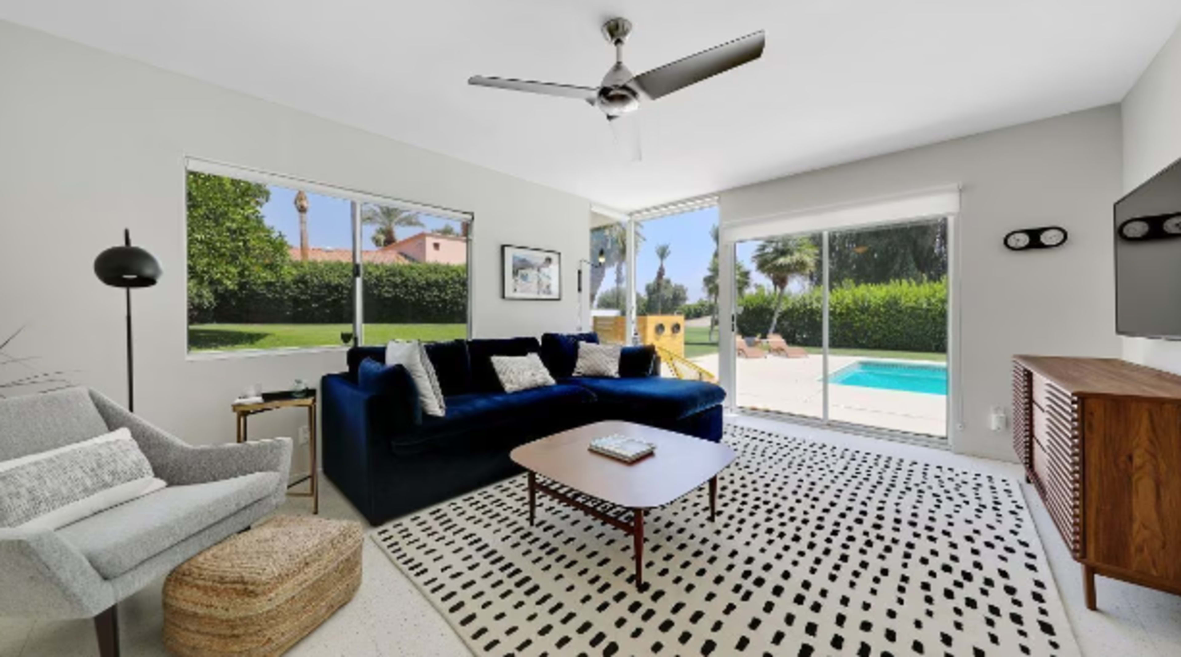 The room features a modern living area with a blue couch, a coffee table, and large windows overlooking a pool and garden.