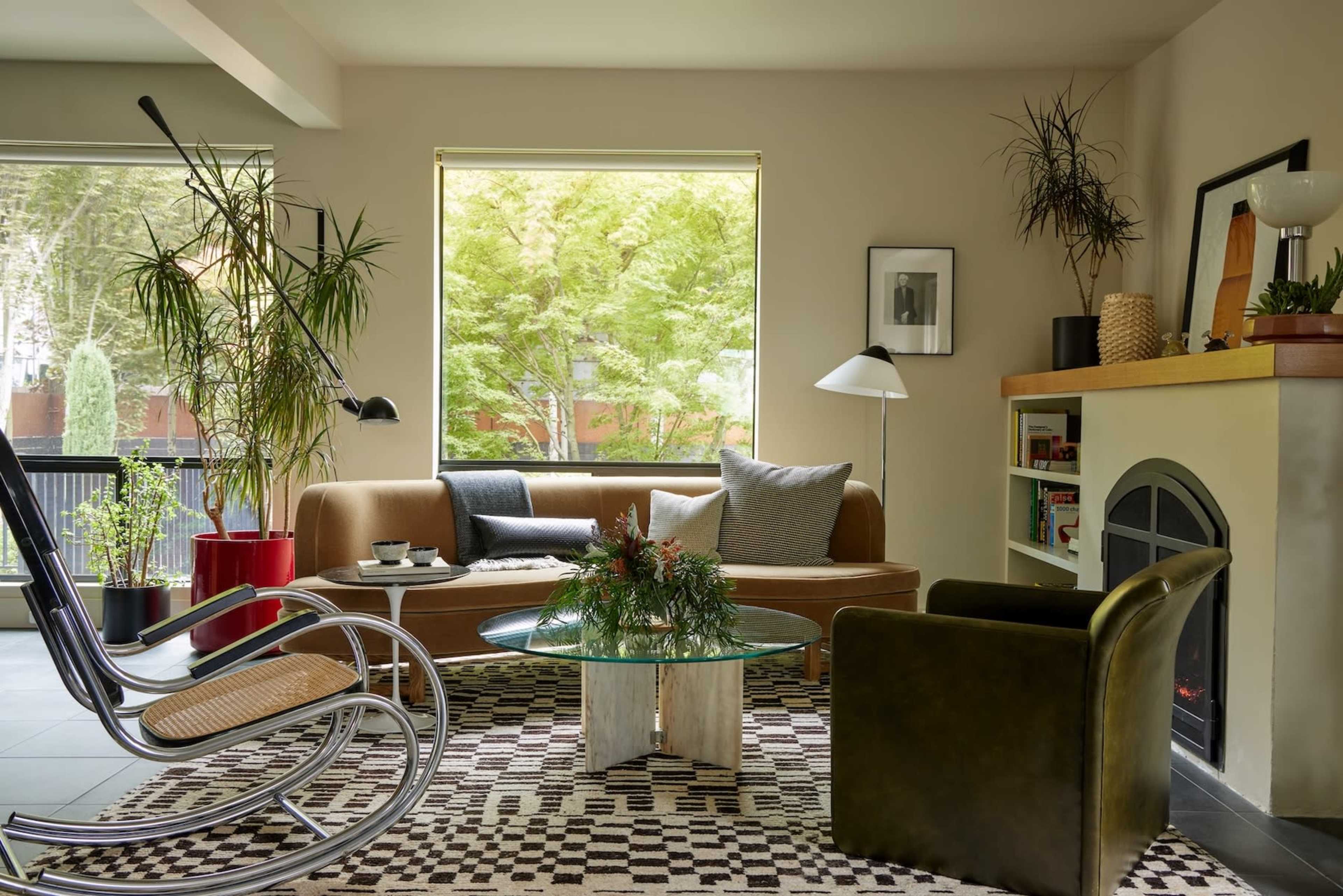 The image shows a modern living room featuring a brown sofa, a glass coffee table, a black armchair, a rocking chair, and large windows with greenery visible outside.