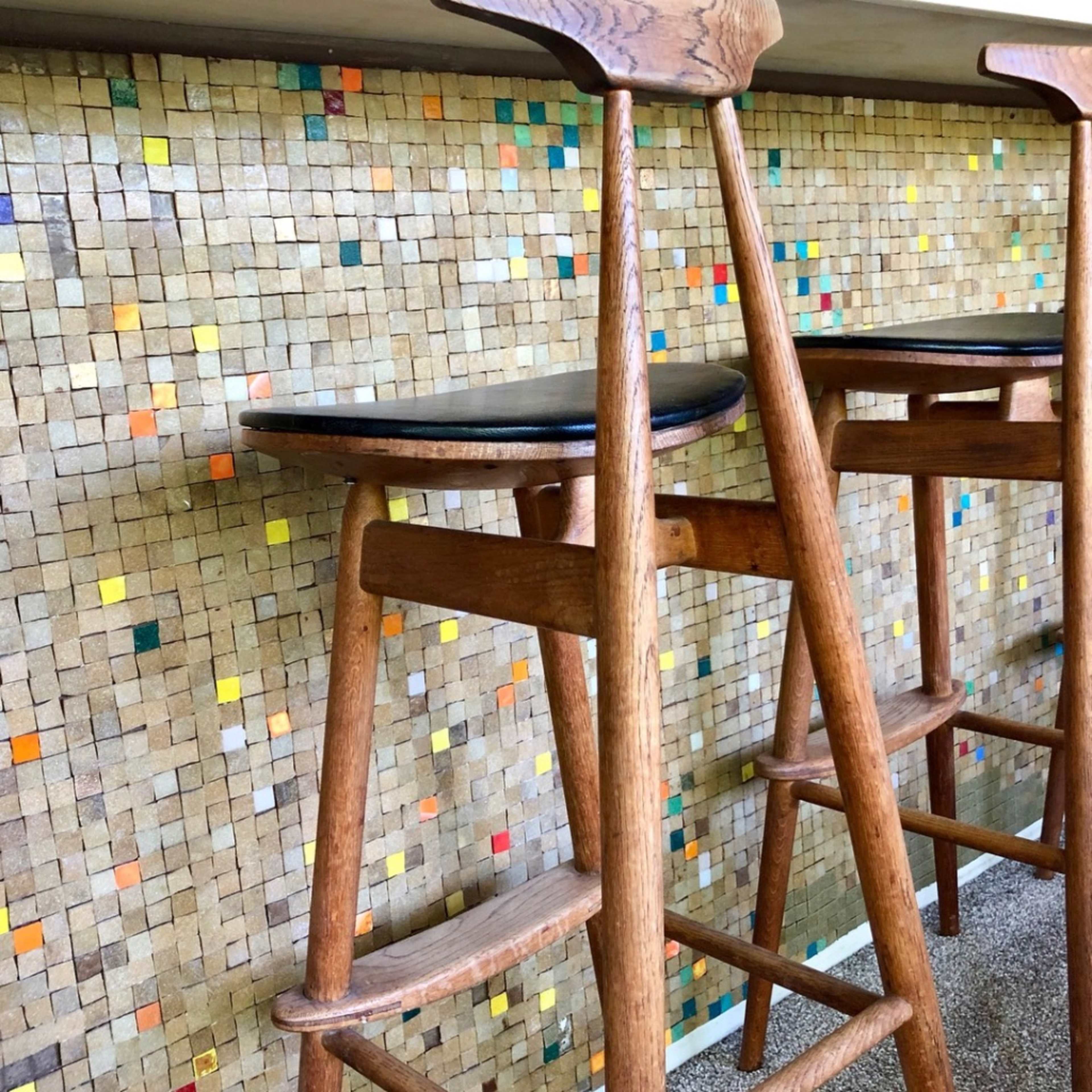Two wooden bar stools are positioned in front of a tiled wall featuring a mosaic of small, square tiles in various colors.