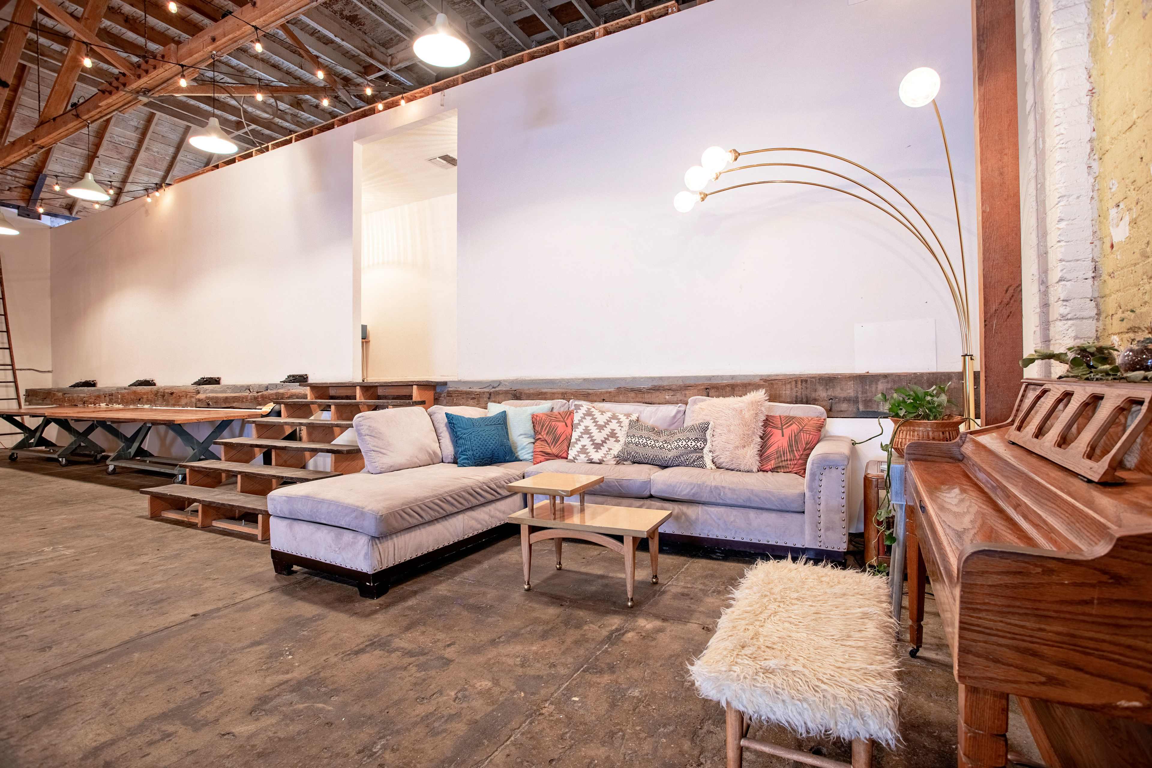 A spacious room featuring a large sectional sofa with decorative pillows, a small table, a piano, and wooden stage-like seating against a white wall.
