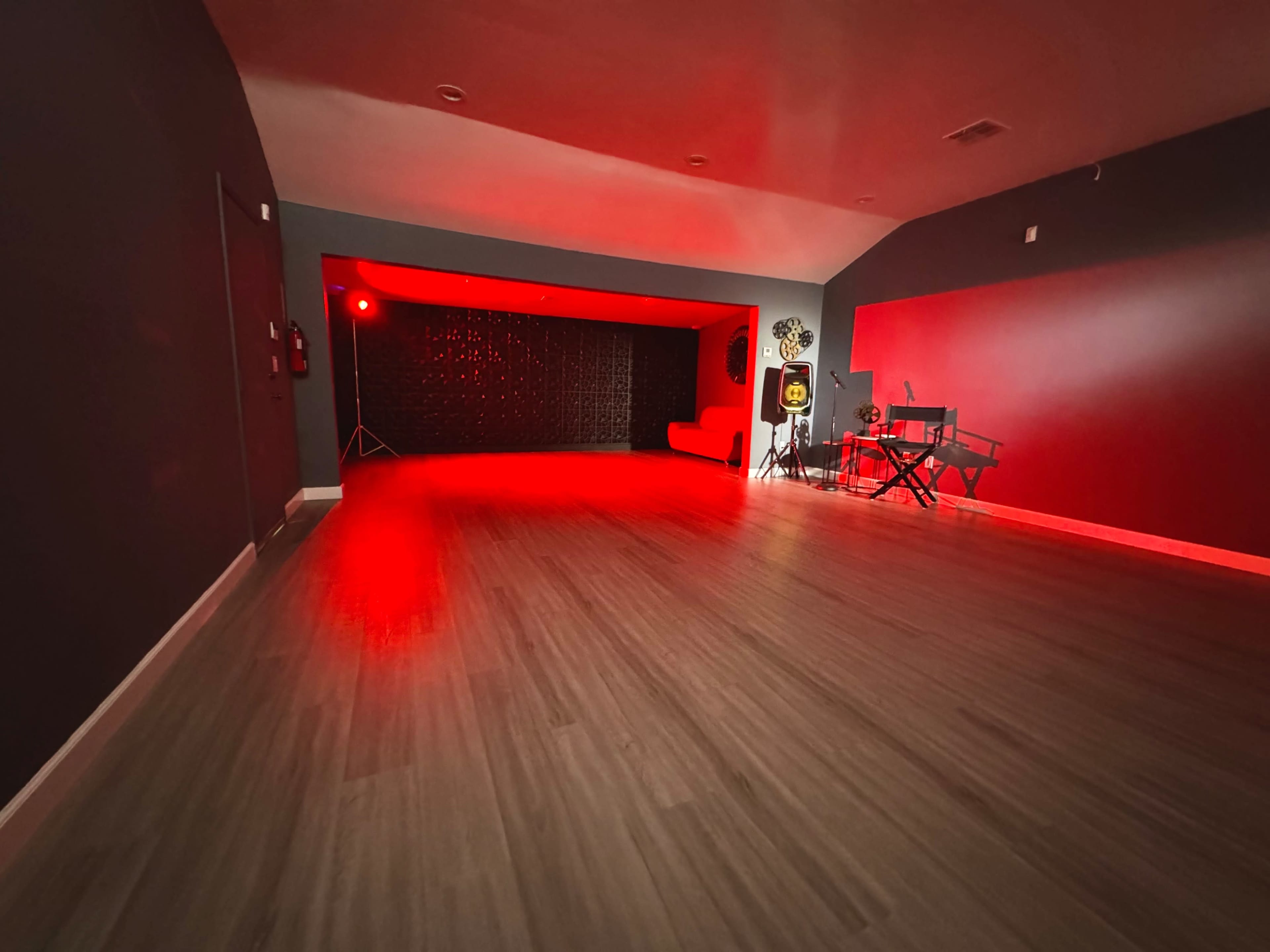 The image shows a spacious room with a wooden floor, dimly lit by red lighting, and featuring a music setup in one corner.