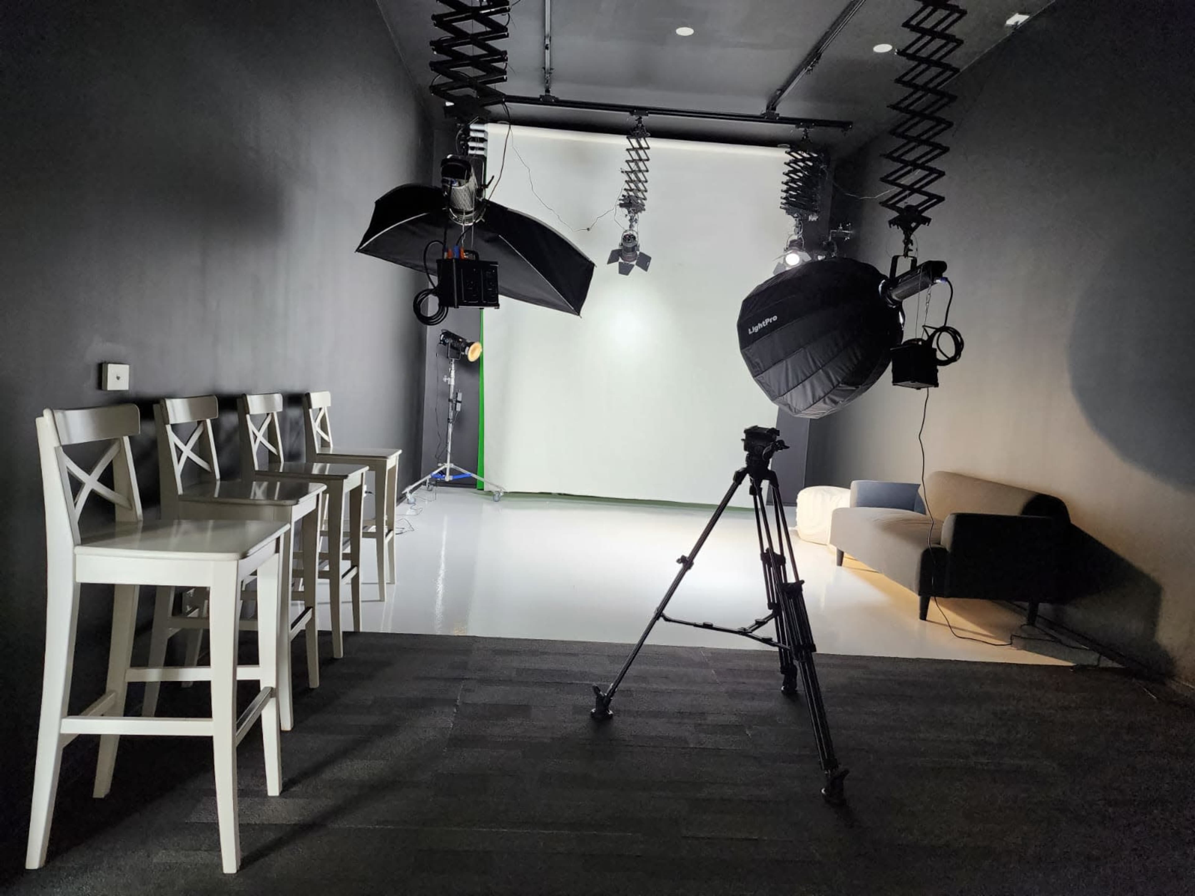 A studio setup featuring white bar stools, a camera on a tripod, and professional lighting equipment, with a green backdrop in the background.
