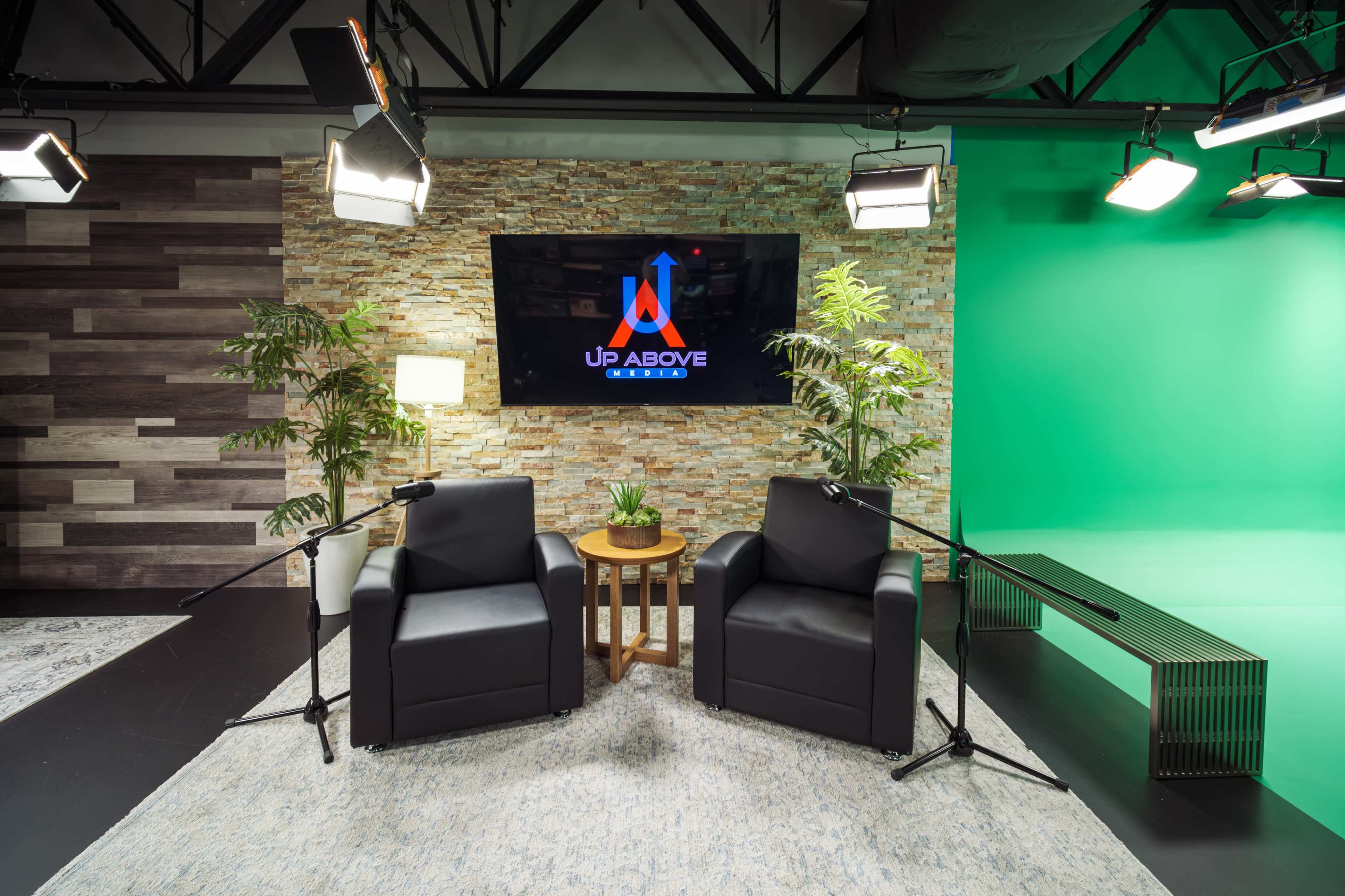 The image shows a studio setup with two black chairs facing a screen displaying the logo "Up Above," surrounded by plants and a green screen backdrop.
