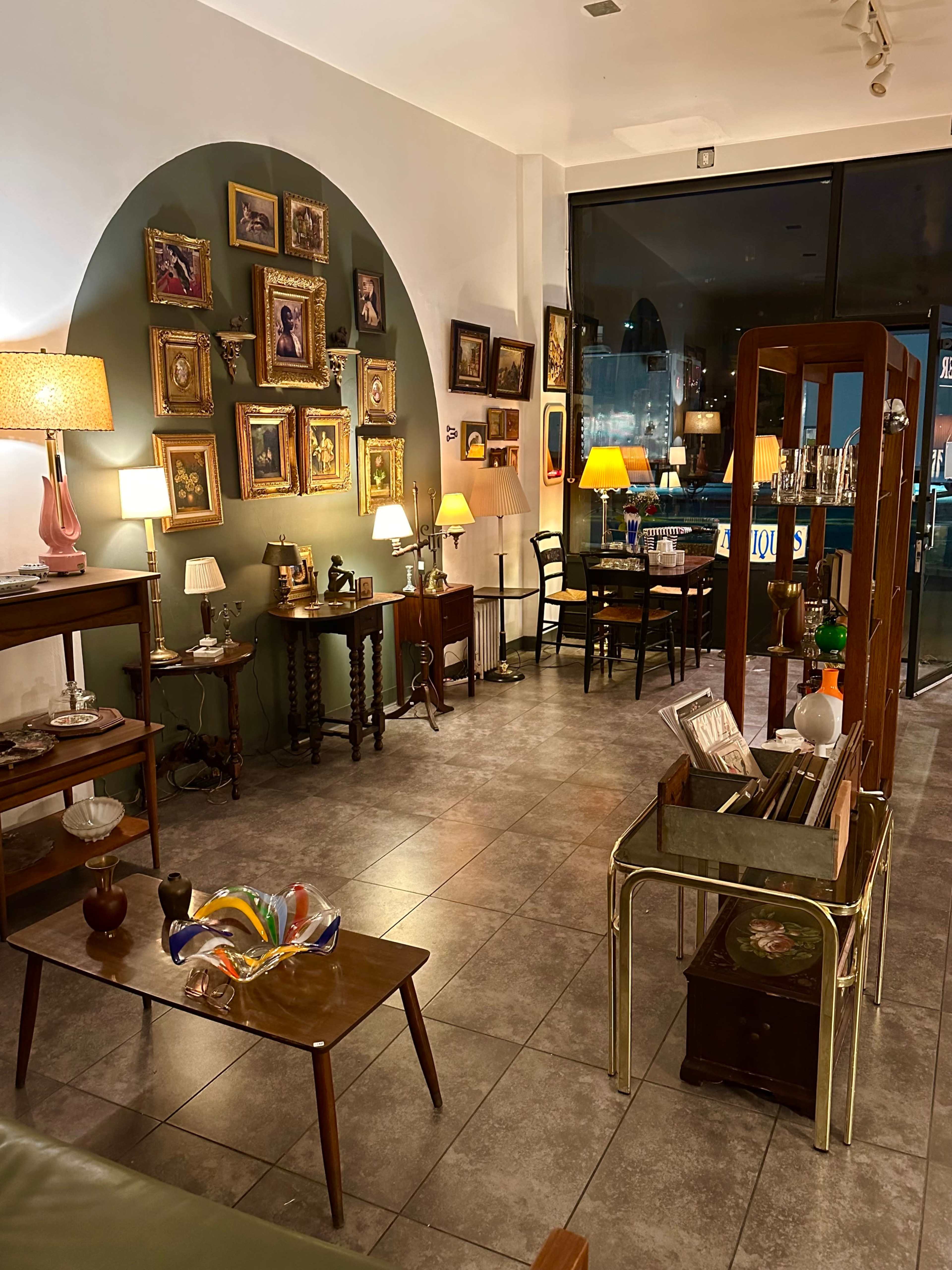 The image shows a cozy antique shop filled with wooden furniture, framed paintings on the walls, and various decorative items arranged on tables and shelves.