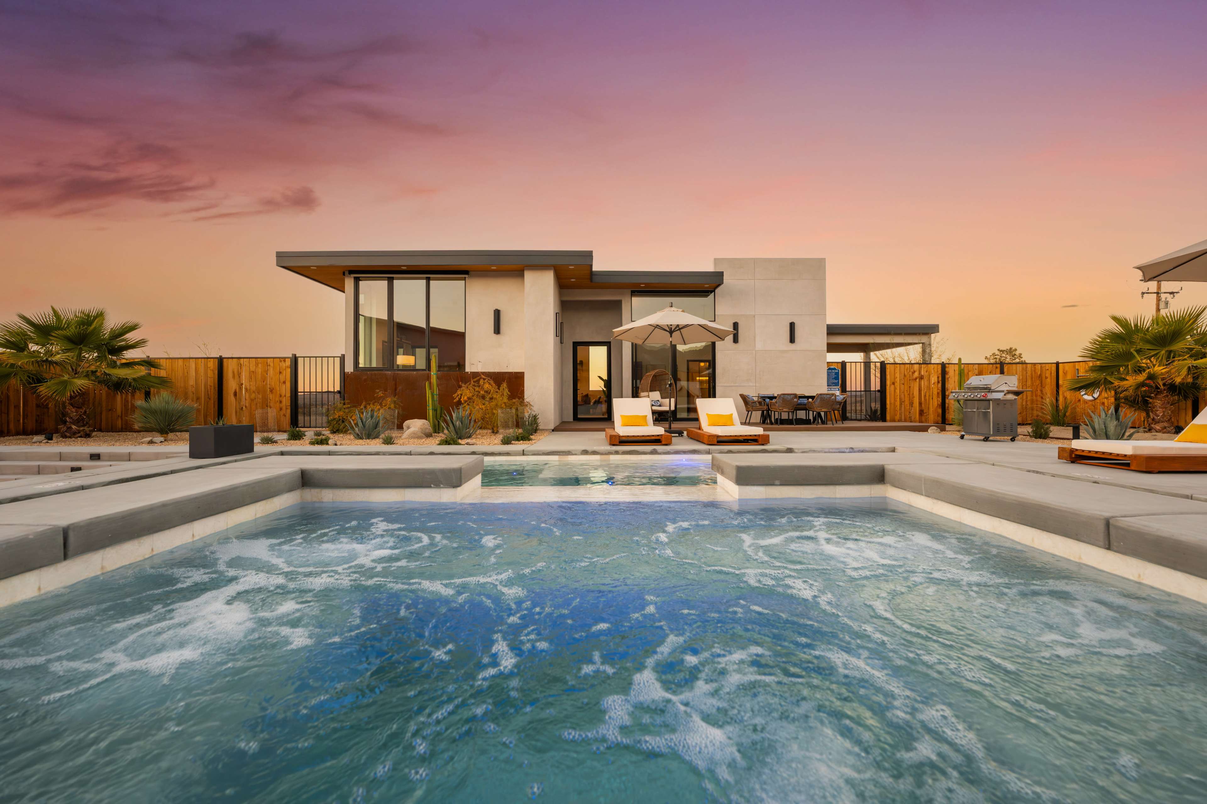 A modern house with a swimming pool and lounge chairs is set against a colorful sunset sky.