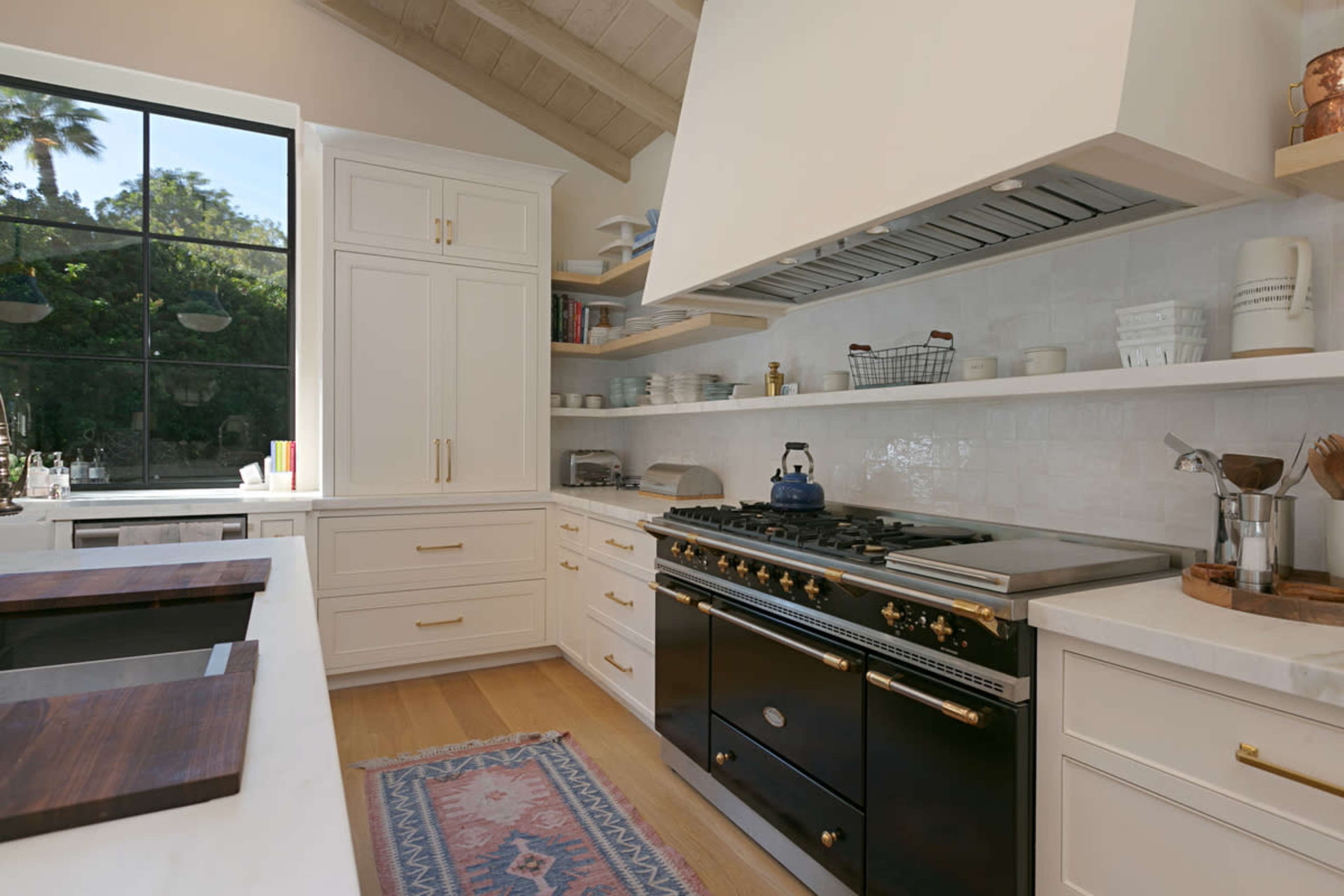 The image shows a modern kitchen featuring white cabinetry, a large black stove, open shelving with dishware, and a window providing natural light.