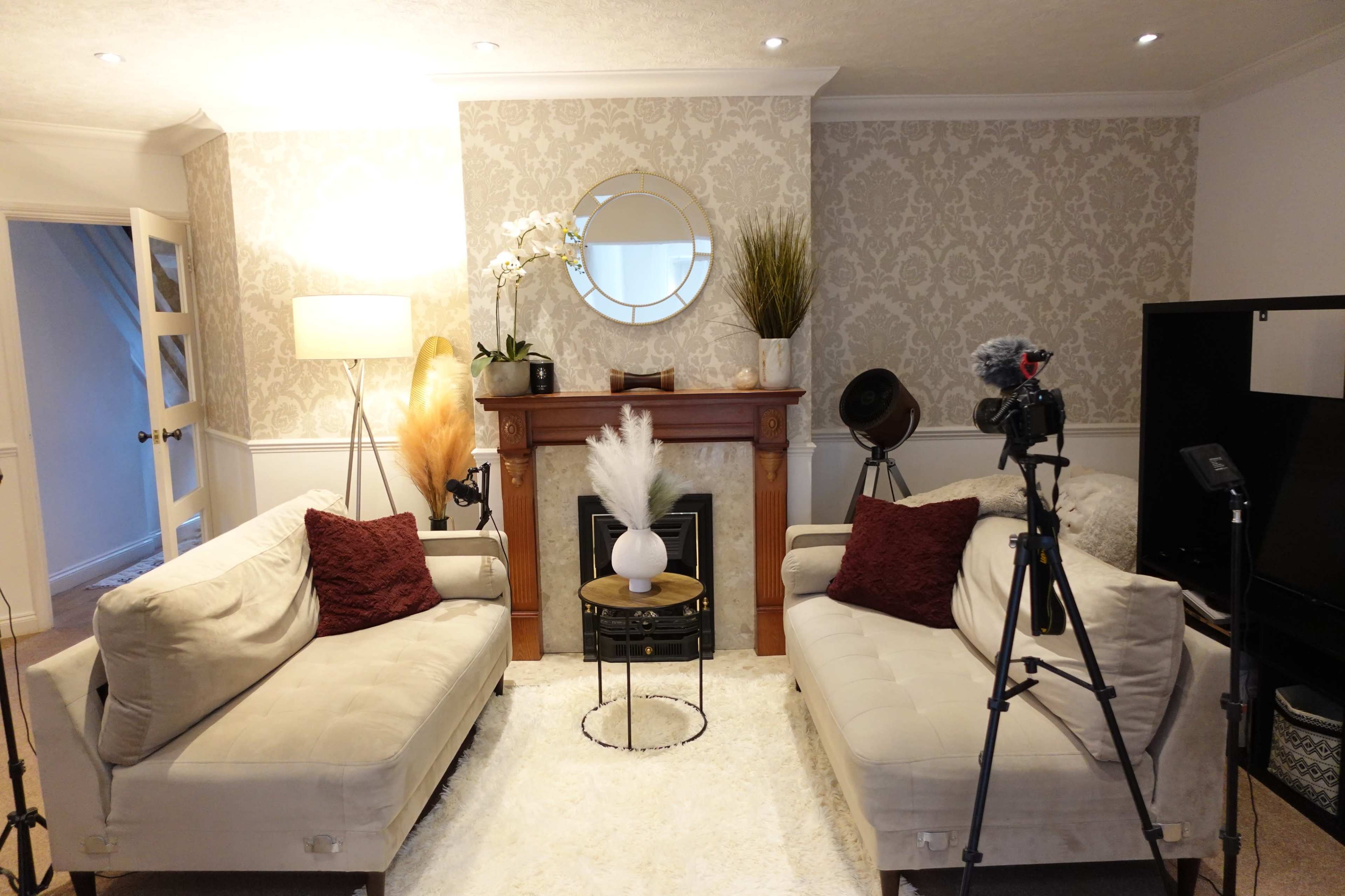 The image shows a well-decorated living room featuring two beige sofas with red pillows, a coffee table, and a fireplace adorned with decorative items.