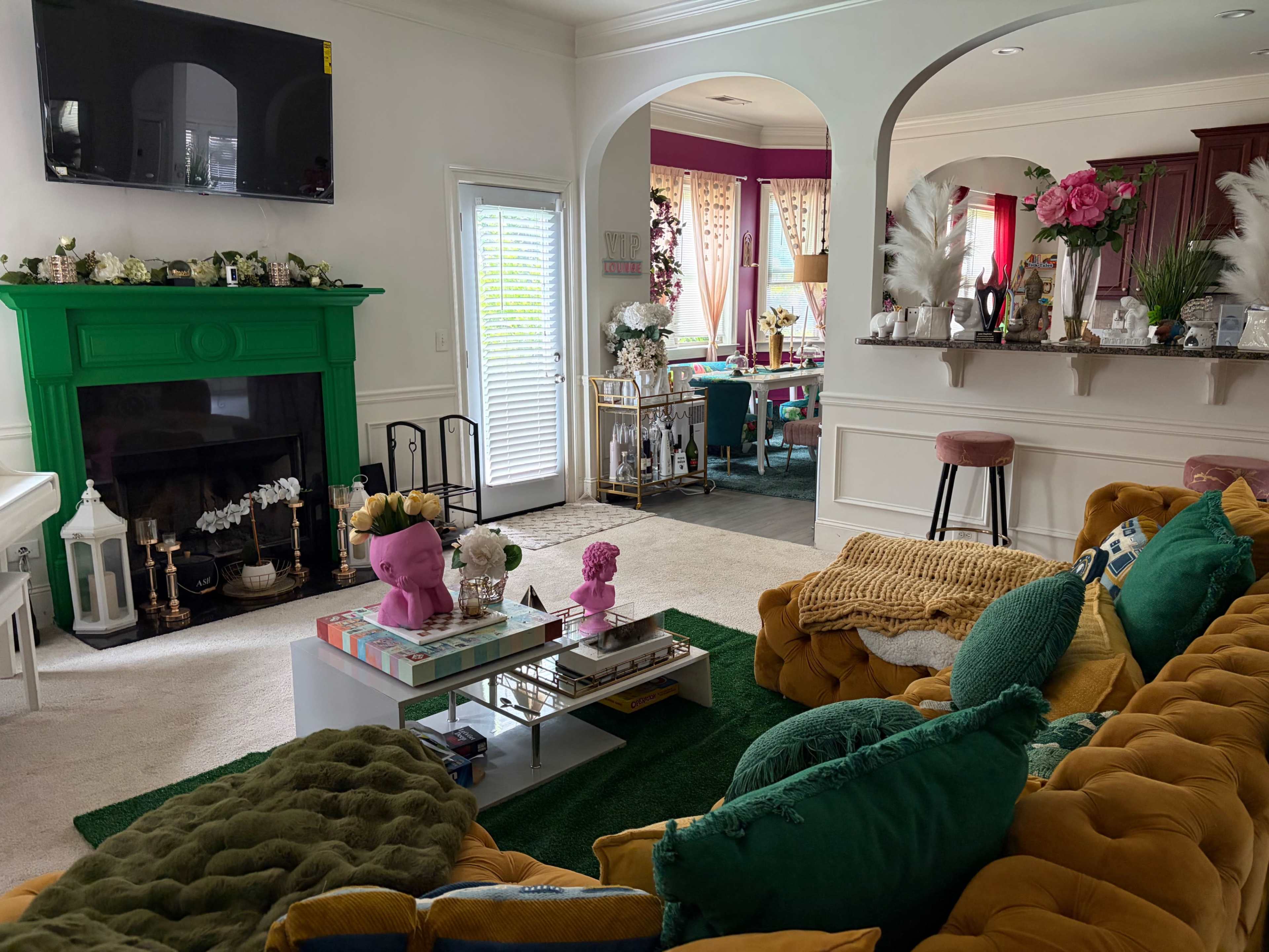 The image shows a brightly decorated living room with a green fireplace, a large yellow sofa, and various decorative items arranged on a coffee table.