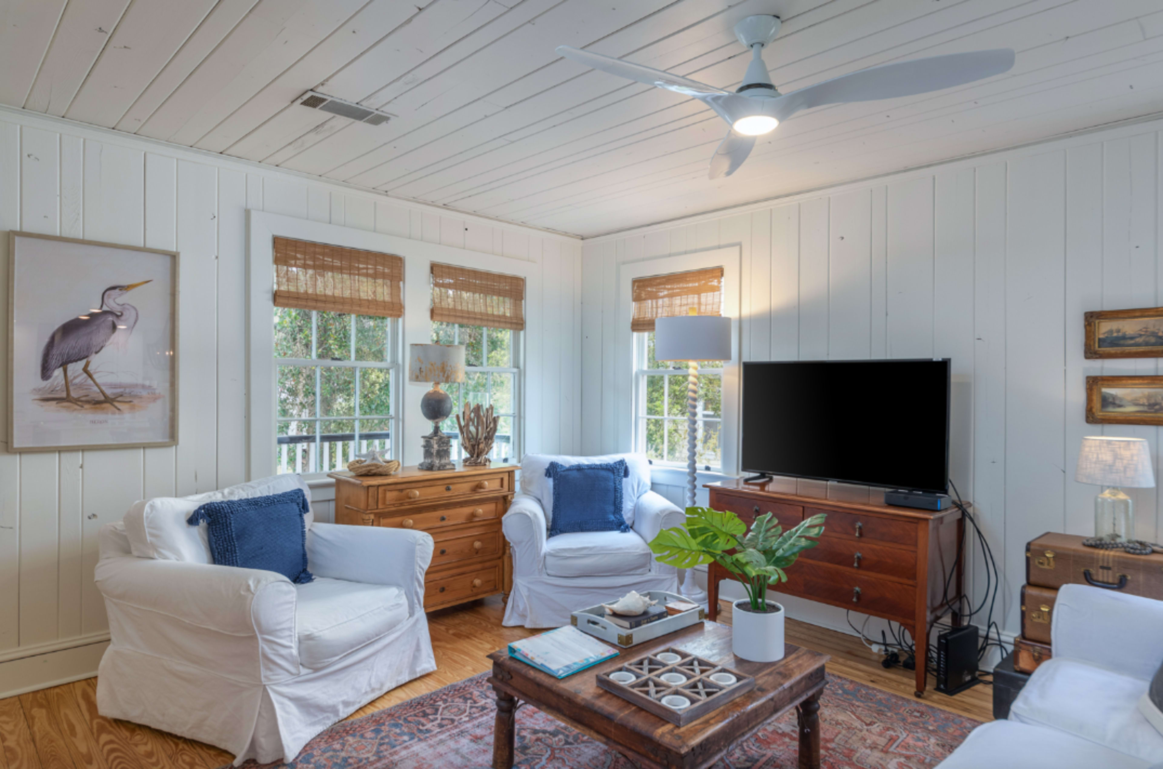 A cozy living room with white walls, a wooden floor, two windows with blinds, and a television on a wooden media console.