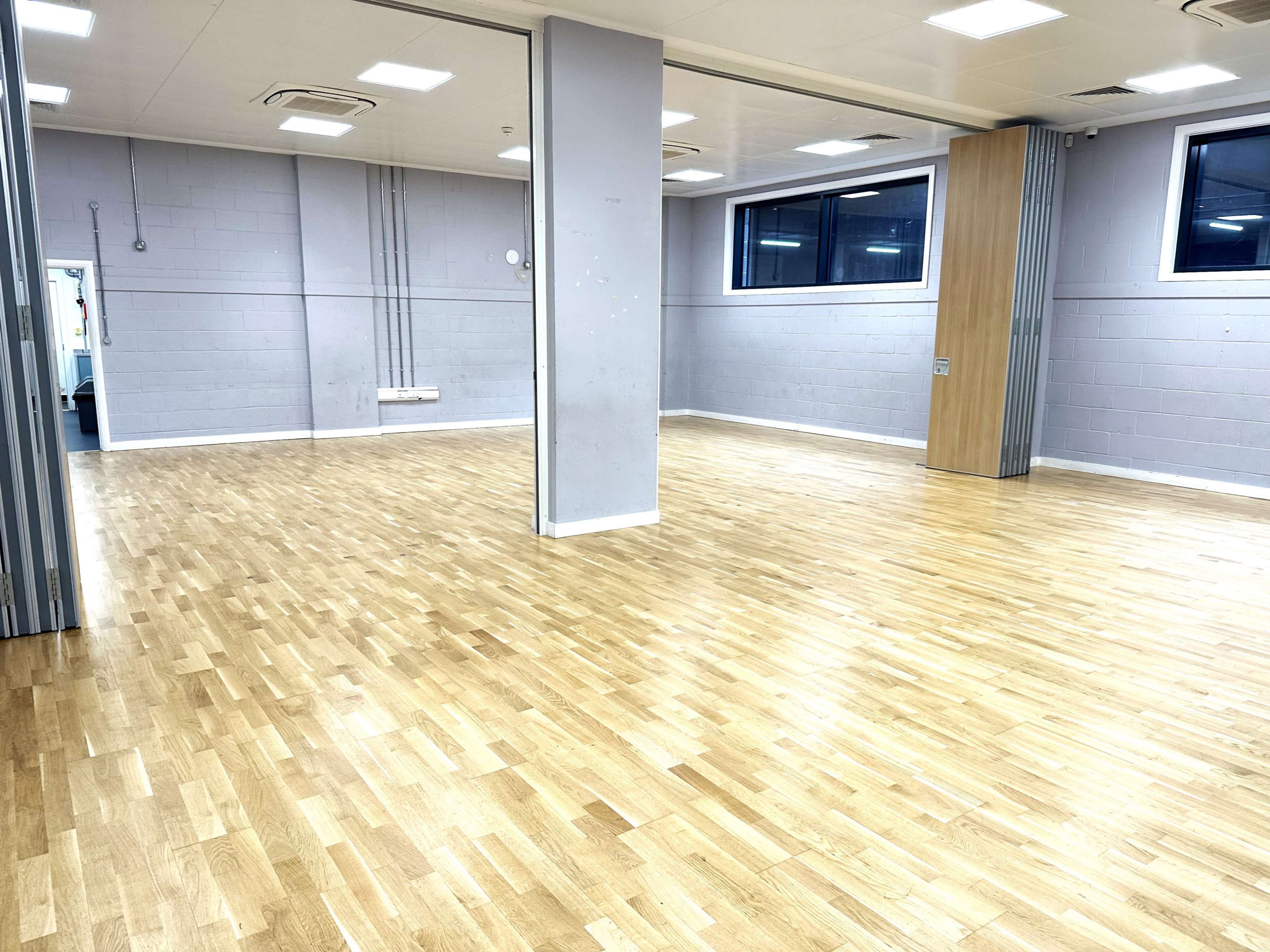 A spacious, empty room with wooden flooring and large windows, featuring gray walls and a central support column.