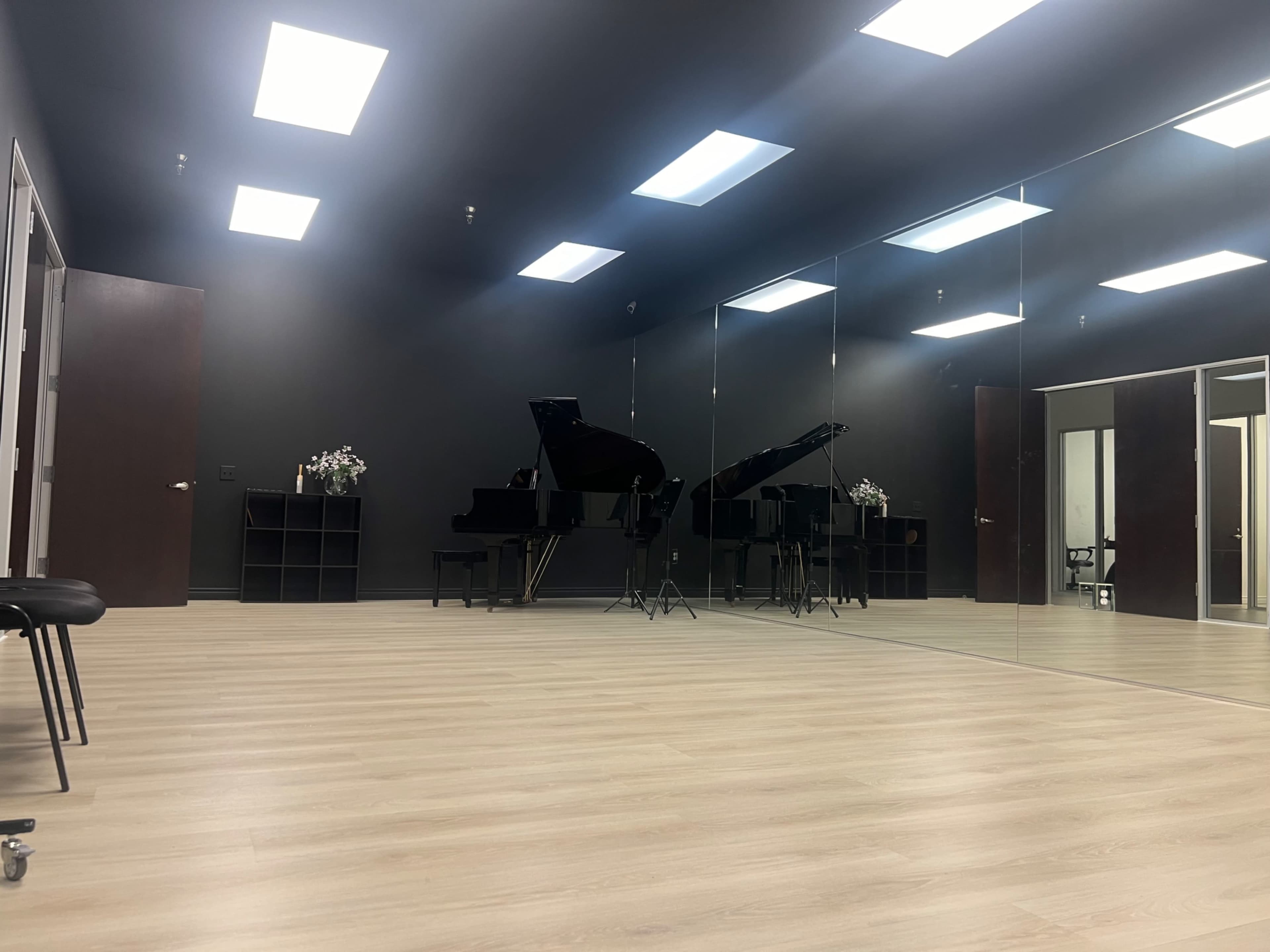 The image shows a music practice room with a black piano, mirrored walls, and wooden flooring under bright overhead lights.