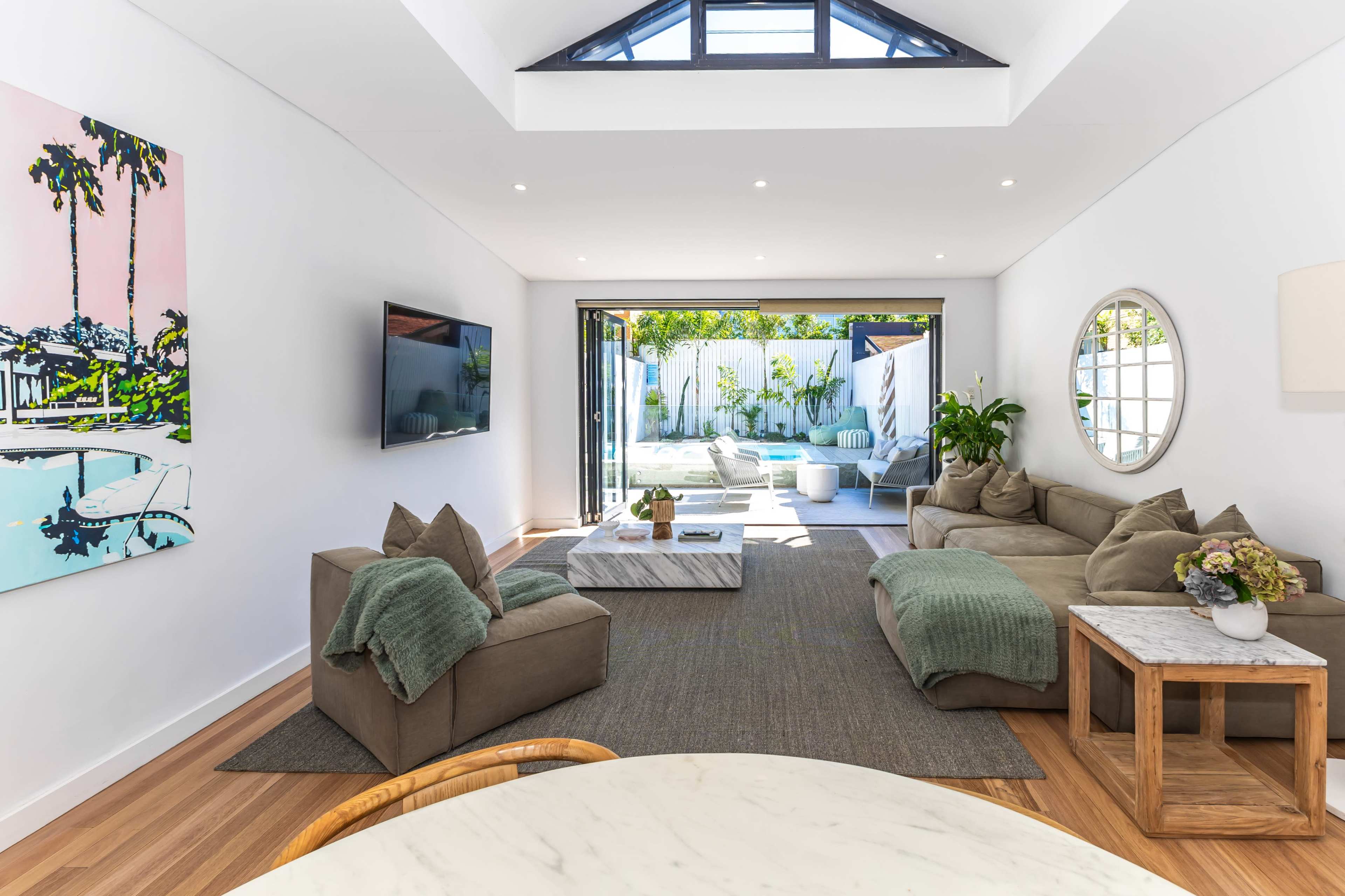 The image shows a modern living room with light walls, two sofas, a round mirror, and large glass doors leading to an outdoor space with a pool.