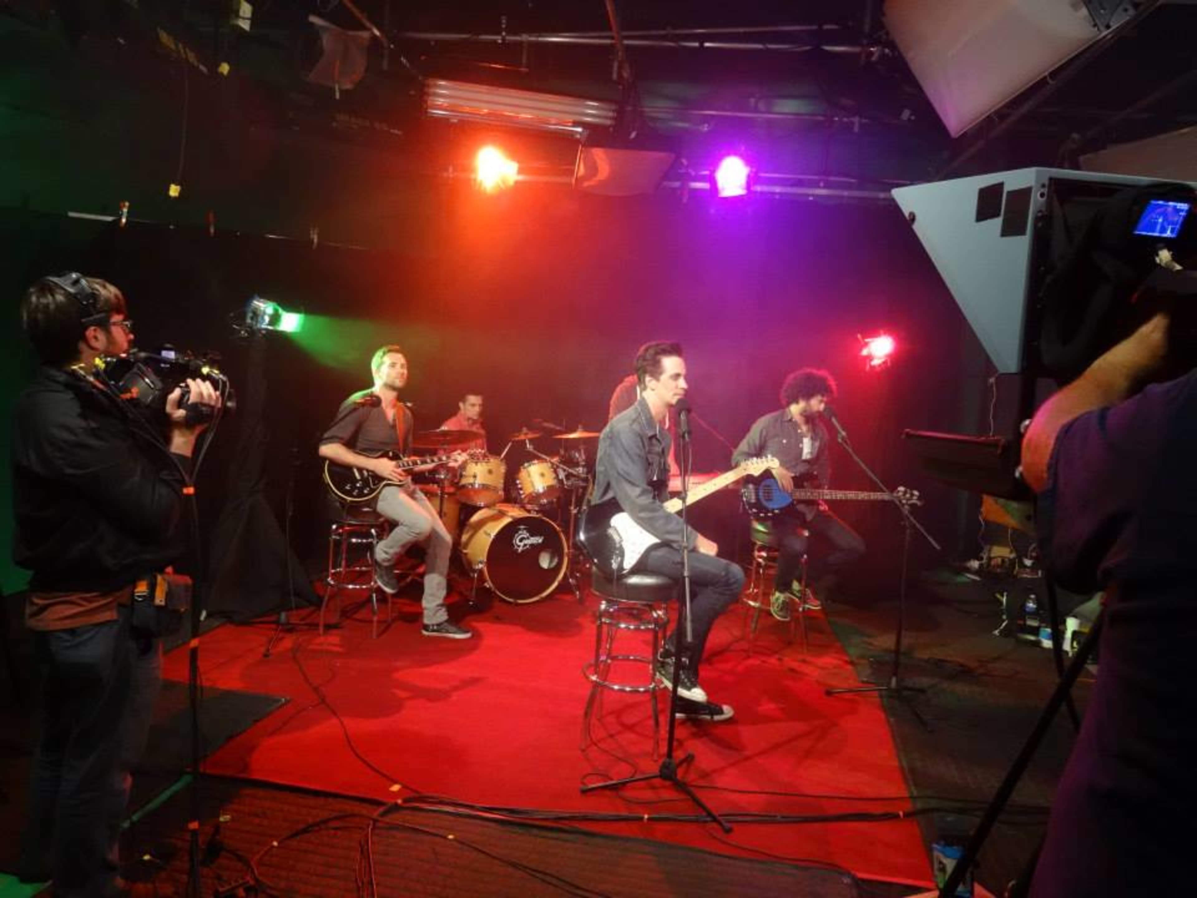 A band performs on a small stage with bright lighting, while a camera crew records the session.