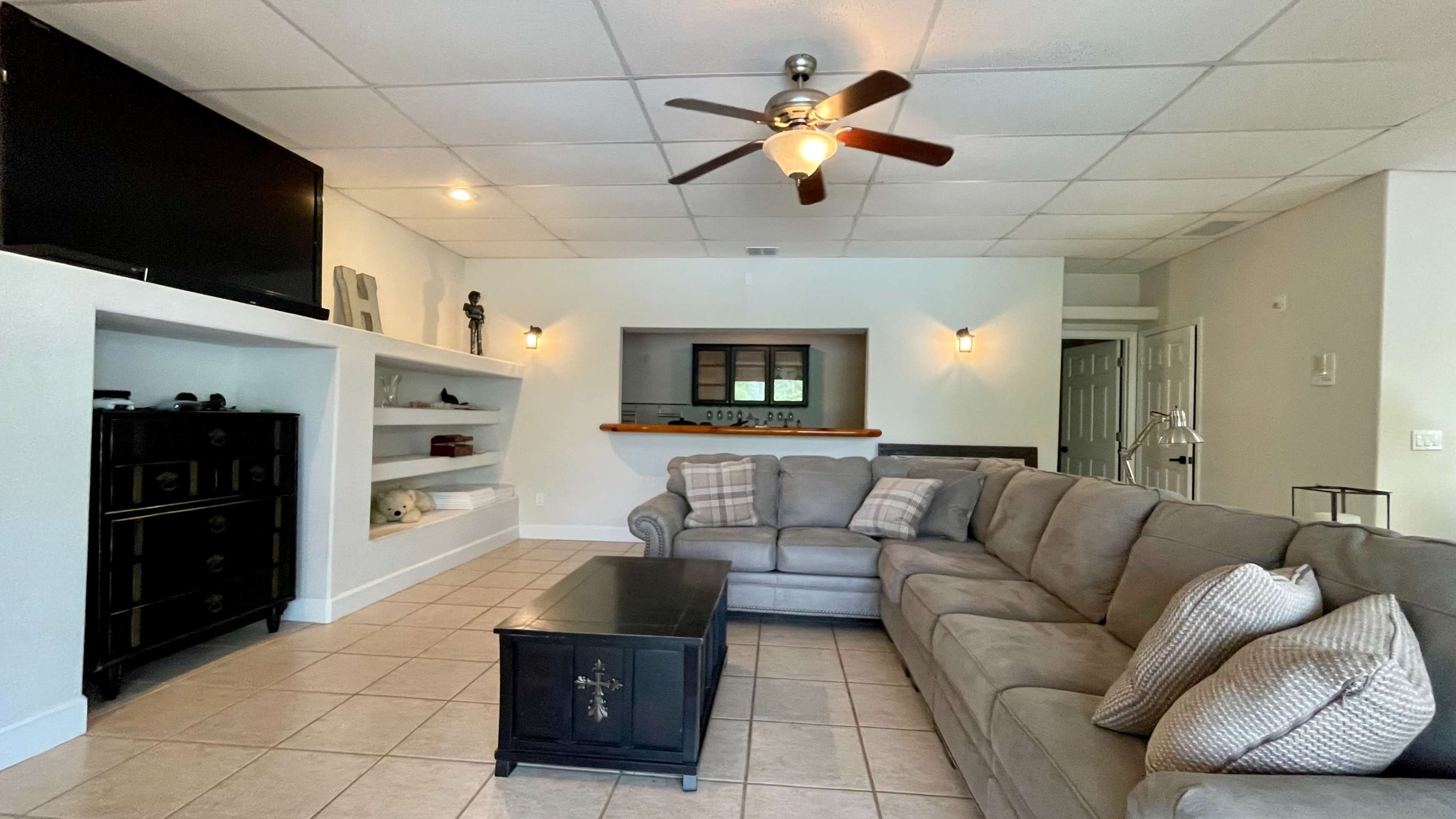 The image shows a living room with a sectional sofa, a coffee table, a flat-screen TV mounted on the wall, and built-in shelves displaying decorative items.