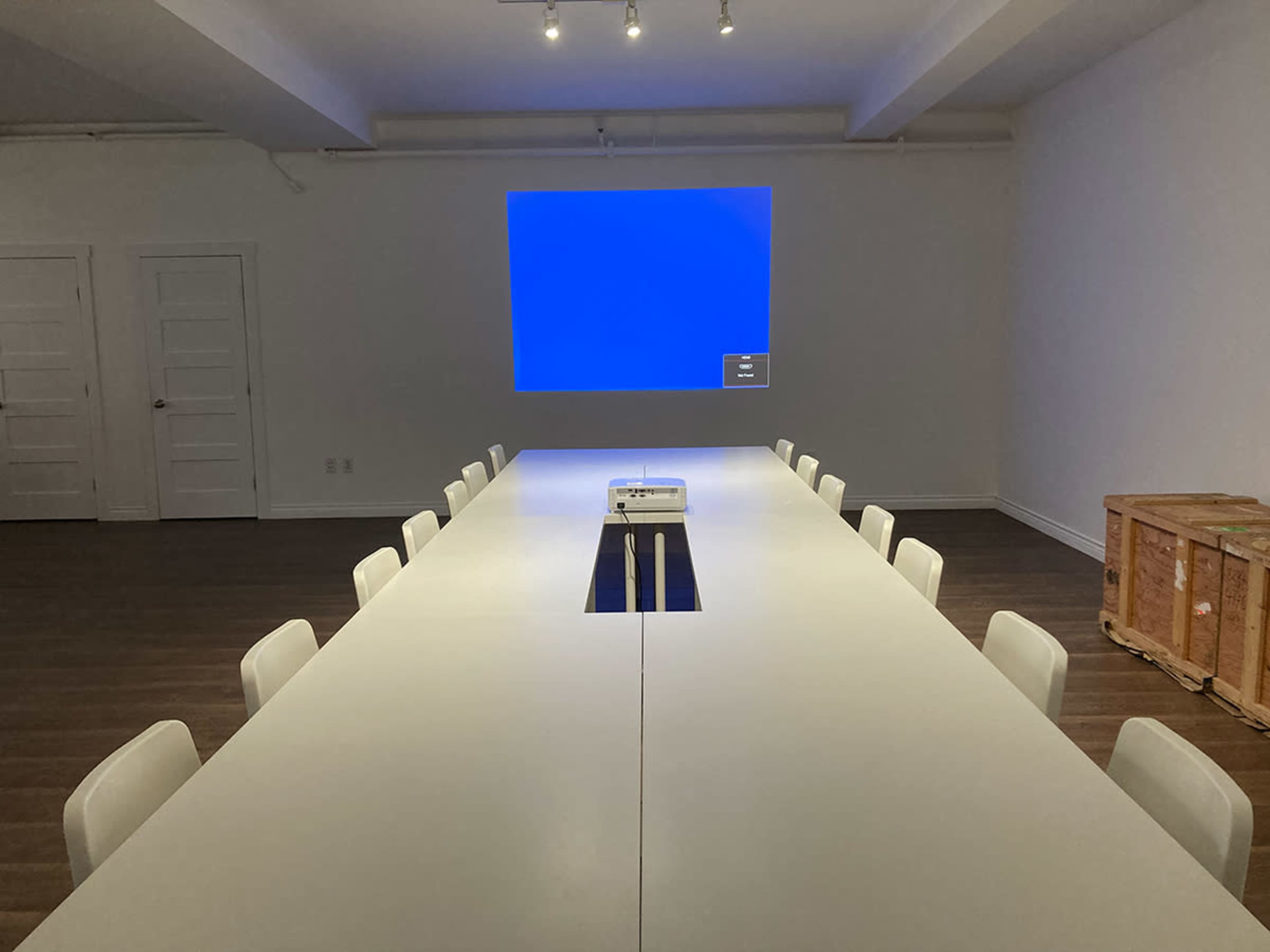 A long, white conference table is set up in a well-lit room with a projector screen displaying a blue background.