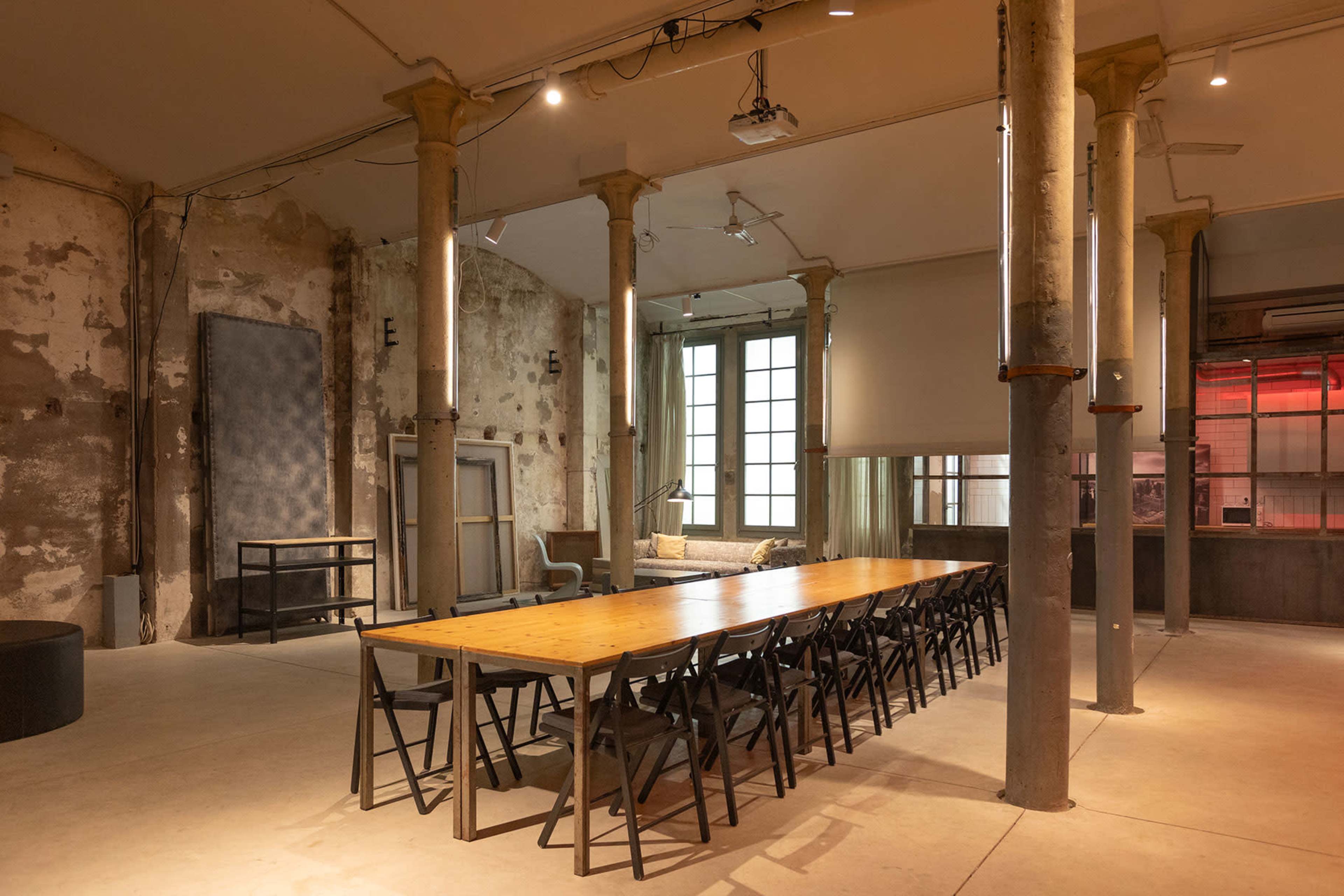 The image shows a spacious, industrial-style meeting room featuring a long wooden table surrounded by black chairs, with exposed columns and large windows letting in natural light.