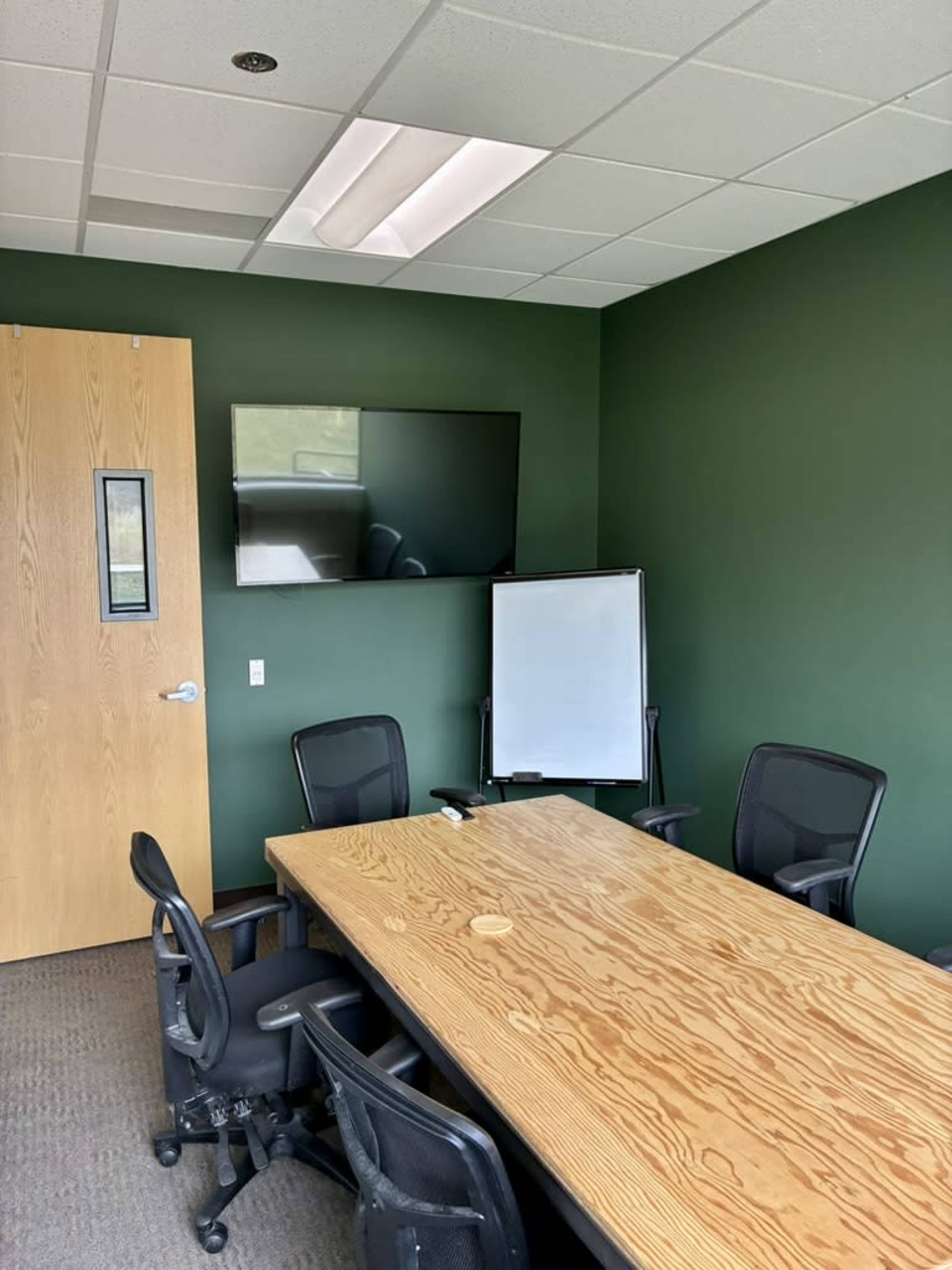 The image shows a conference room with a wooden table, several black chairs, a whiteboard, and a wall-mounted television in a room with green walls.