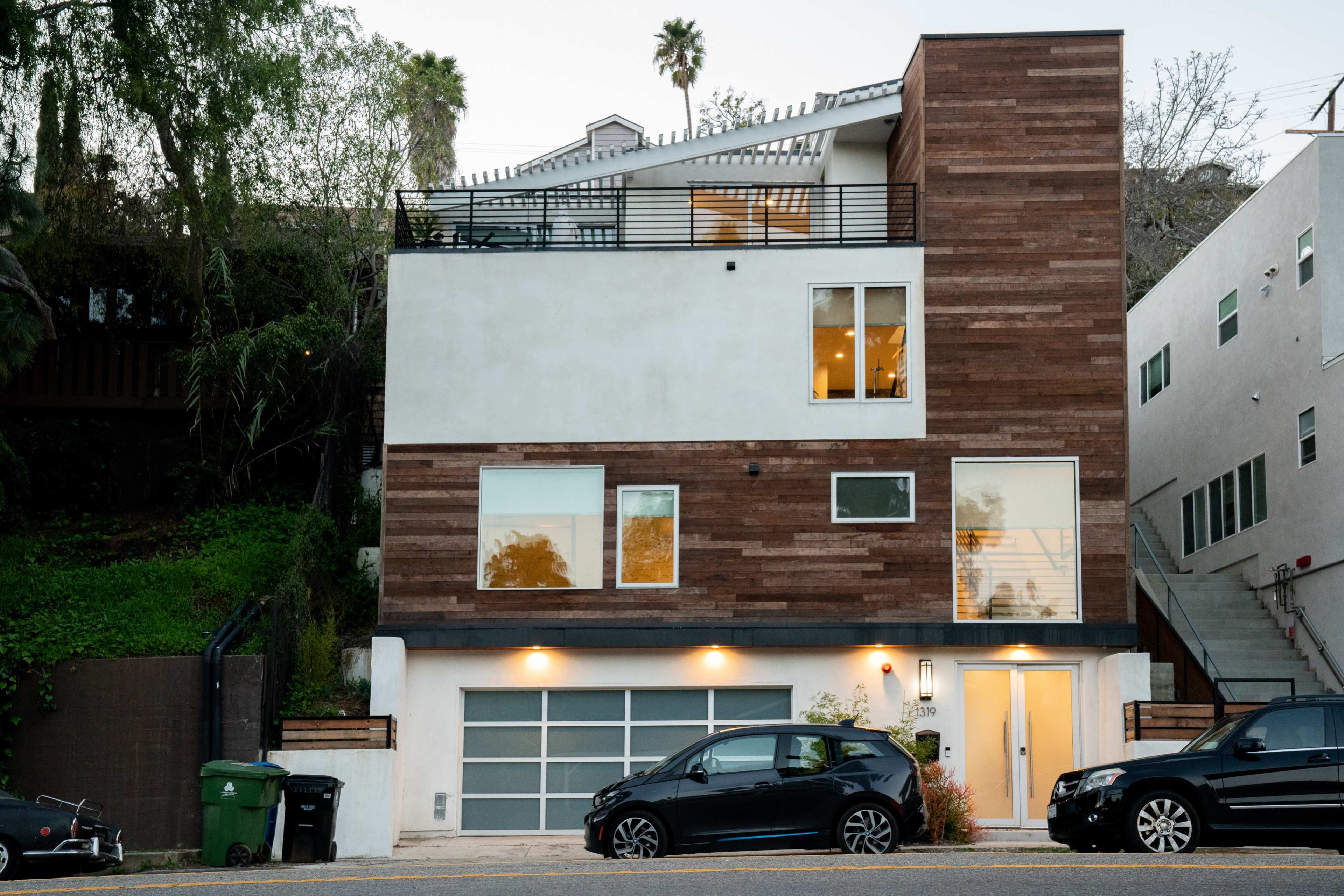 A modern two-story house with a wooden facade and large windows, located on a hillside street with parked cars in front.