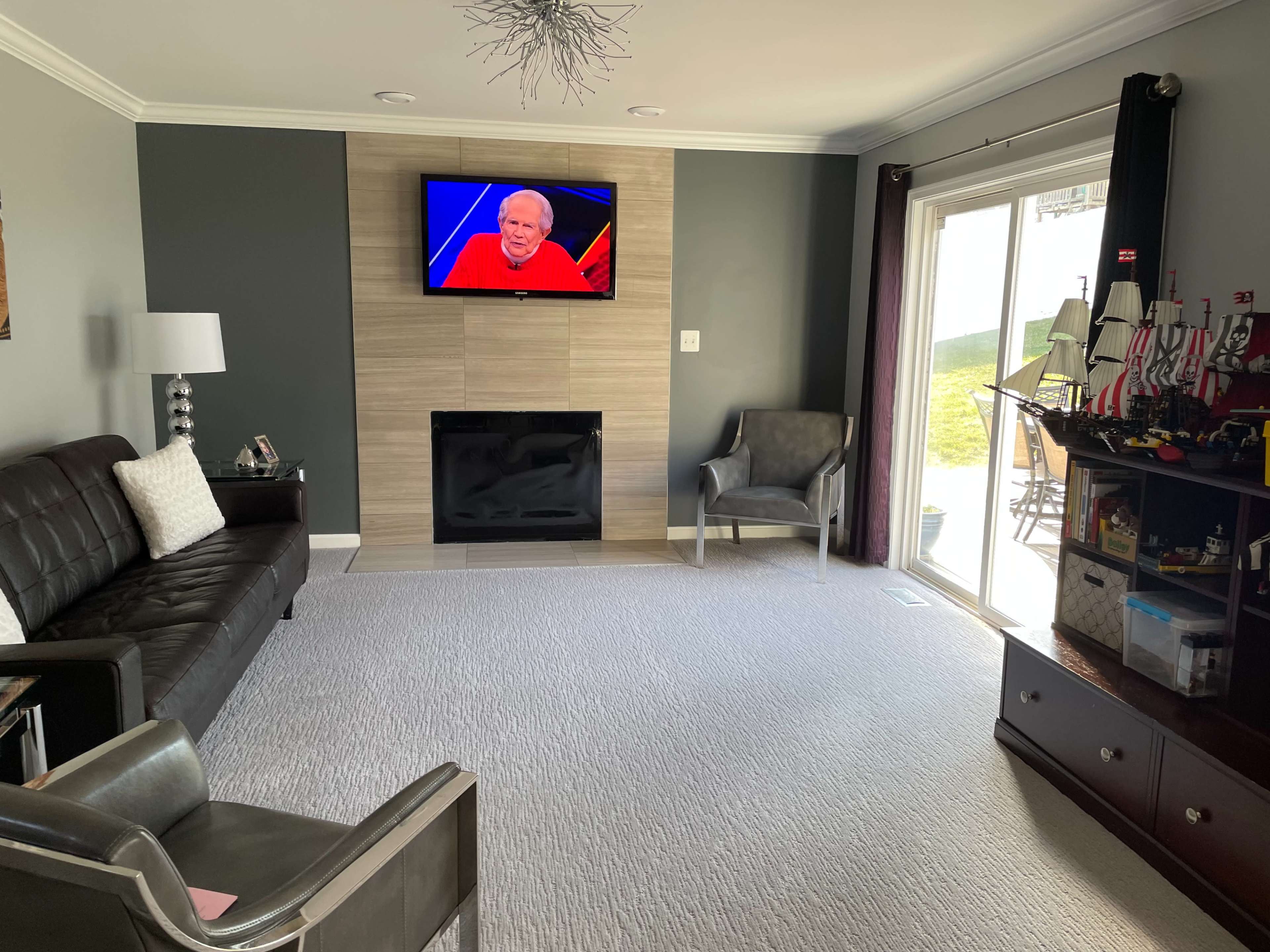 A living room with a dark couch, two chairs, a television mounted on the wall, and a large window leading to an outside view.