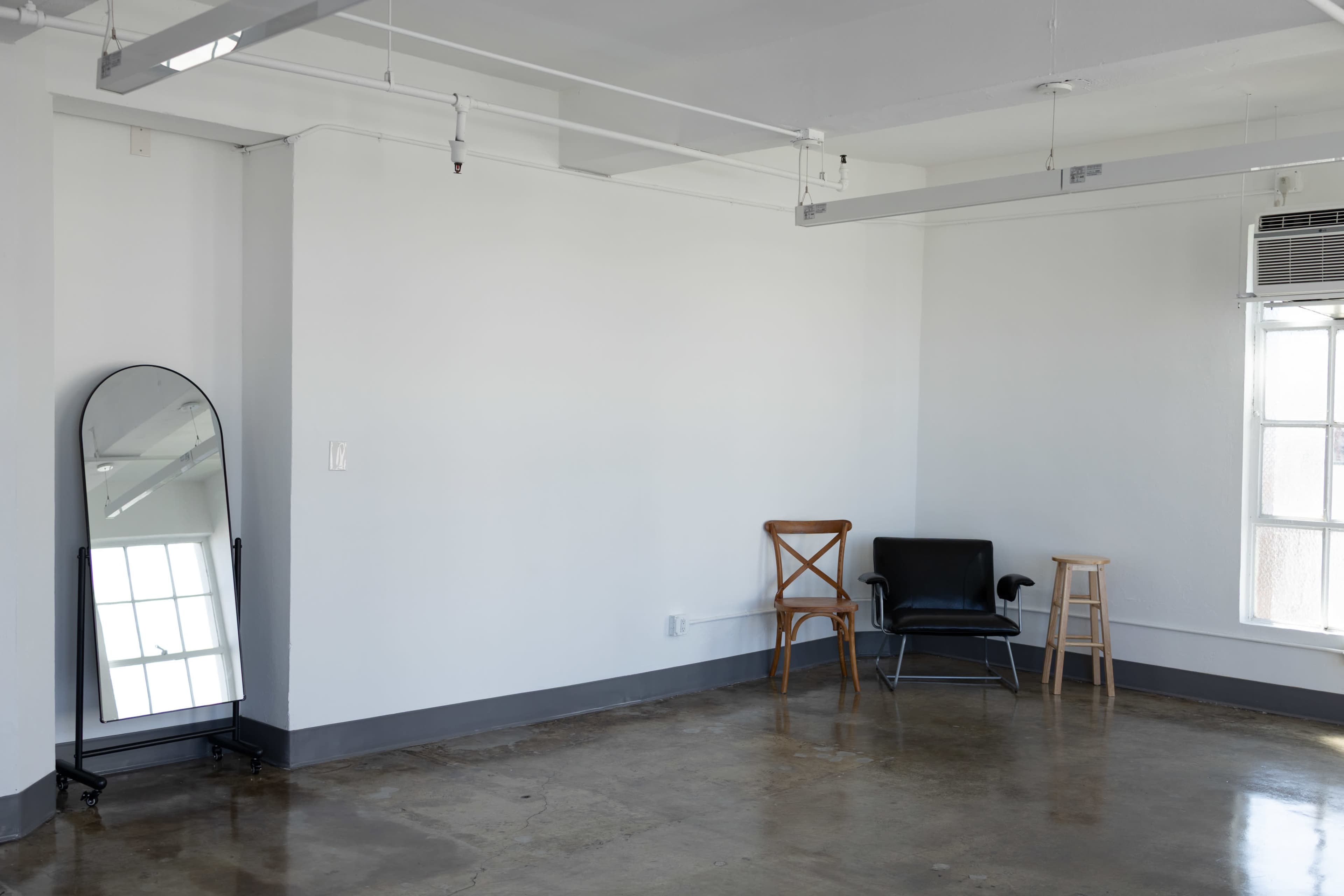 The image shows a minimalist interior space featuring a full-length mirror, a black chair, a wooden stool, and a large window.