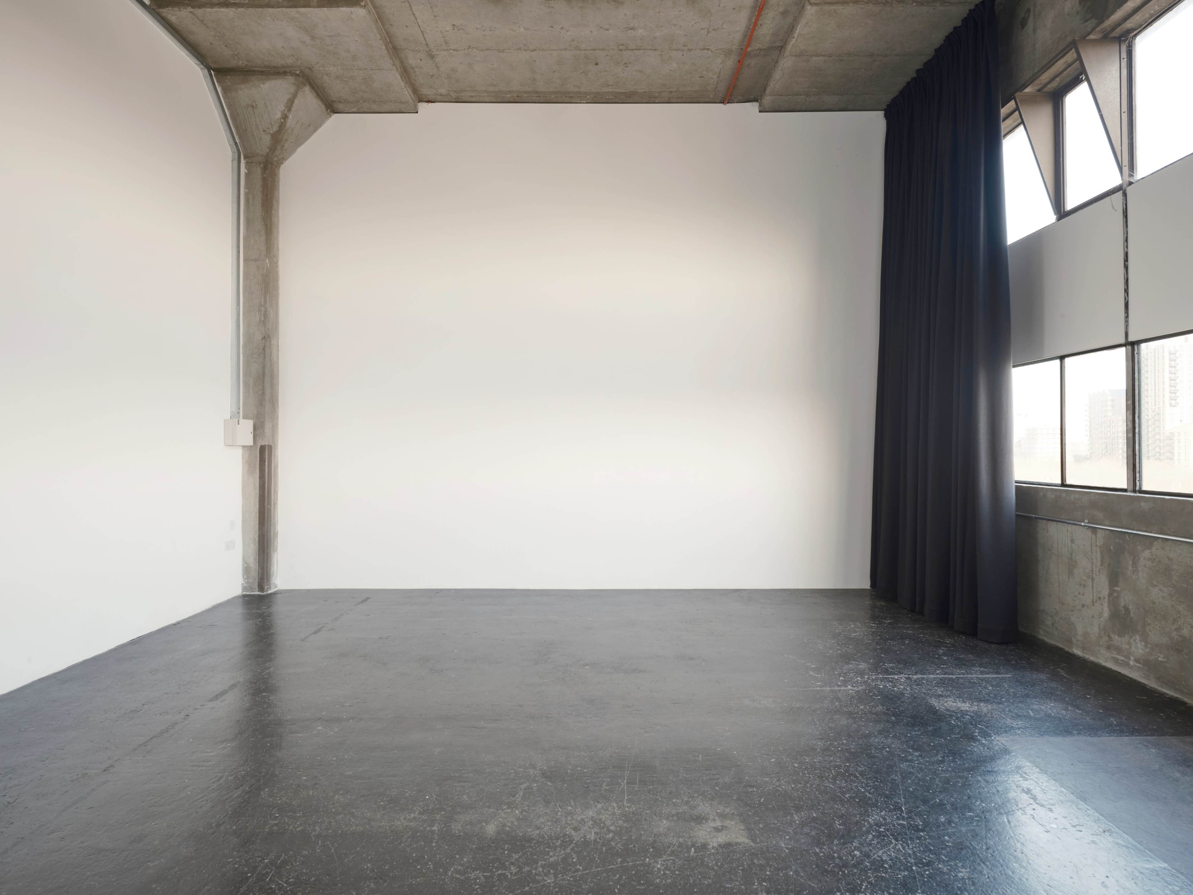 An empty industrial-style room with concrete walls, a dark floor, and large windows covered by a black curtain.
