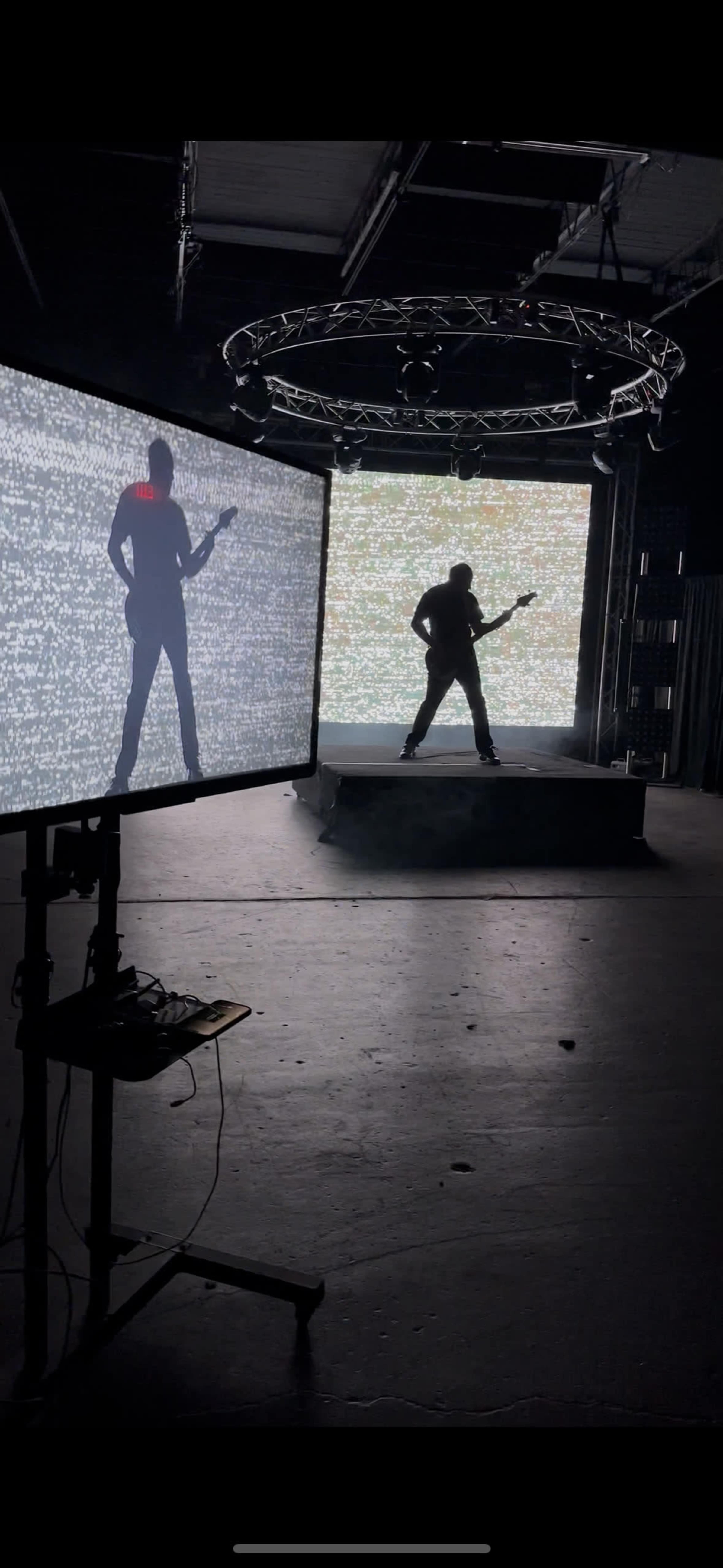 A guitarist performs on a stage while silhouetted against screens displaying static-like visuals.