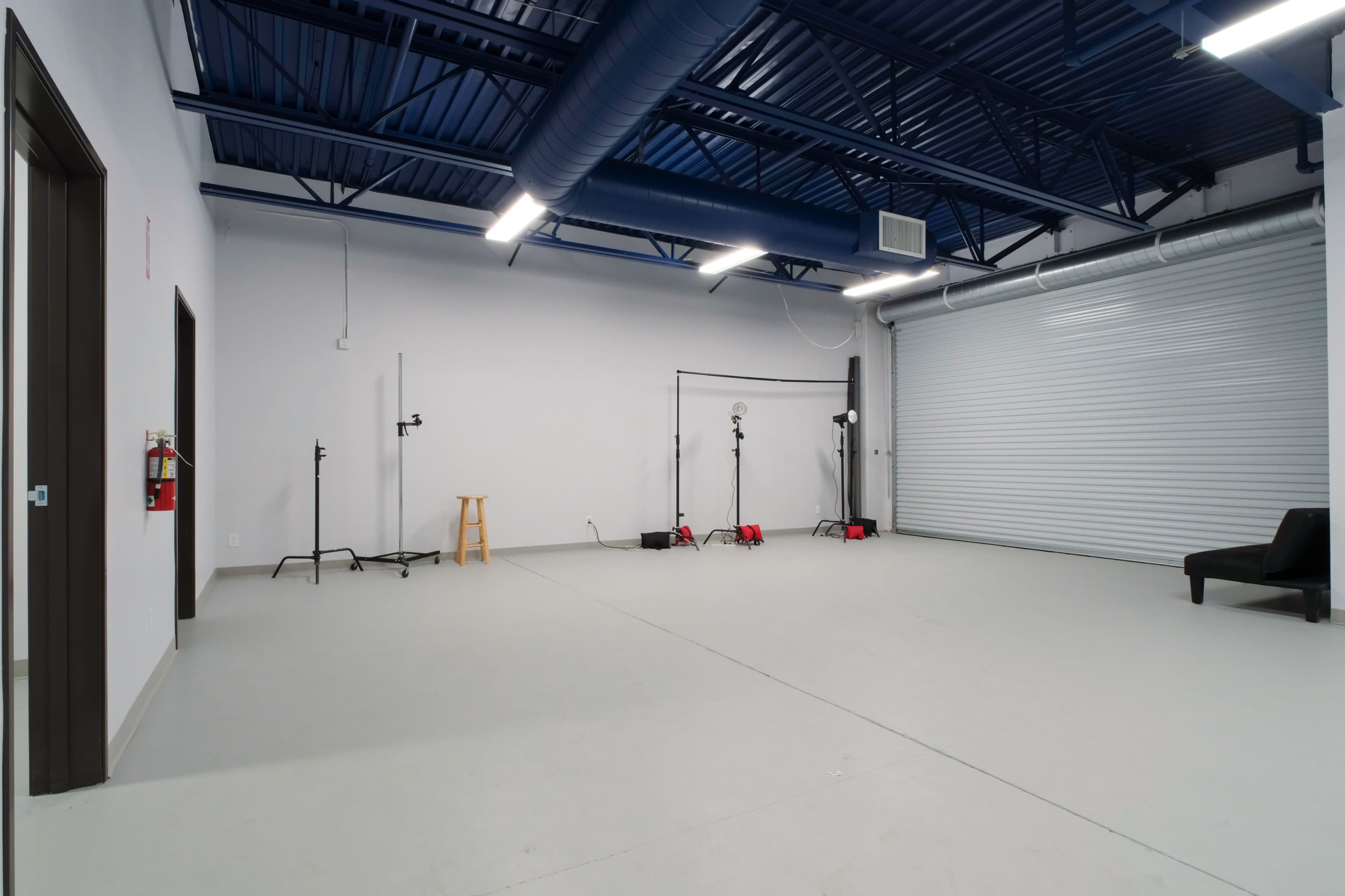 The image shows a spacious, empty studio room with grey flooring, two light stands, and a black chair against a wall.