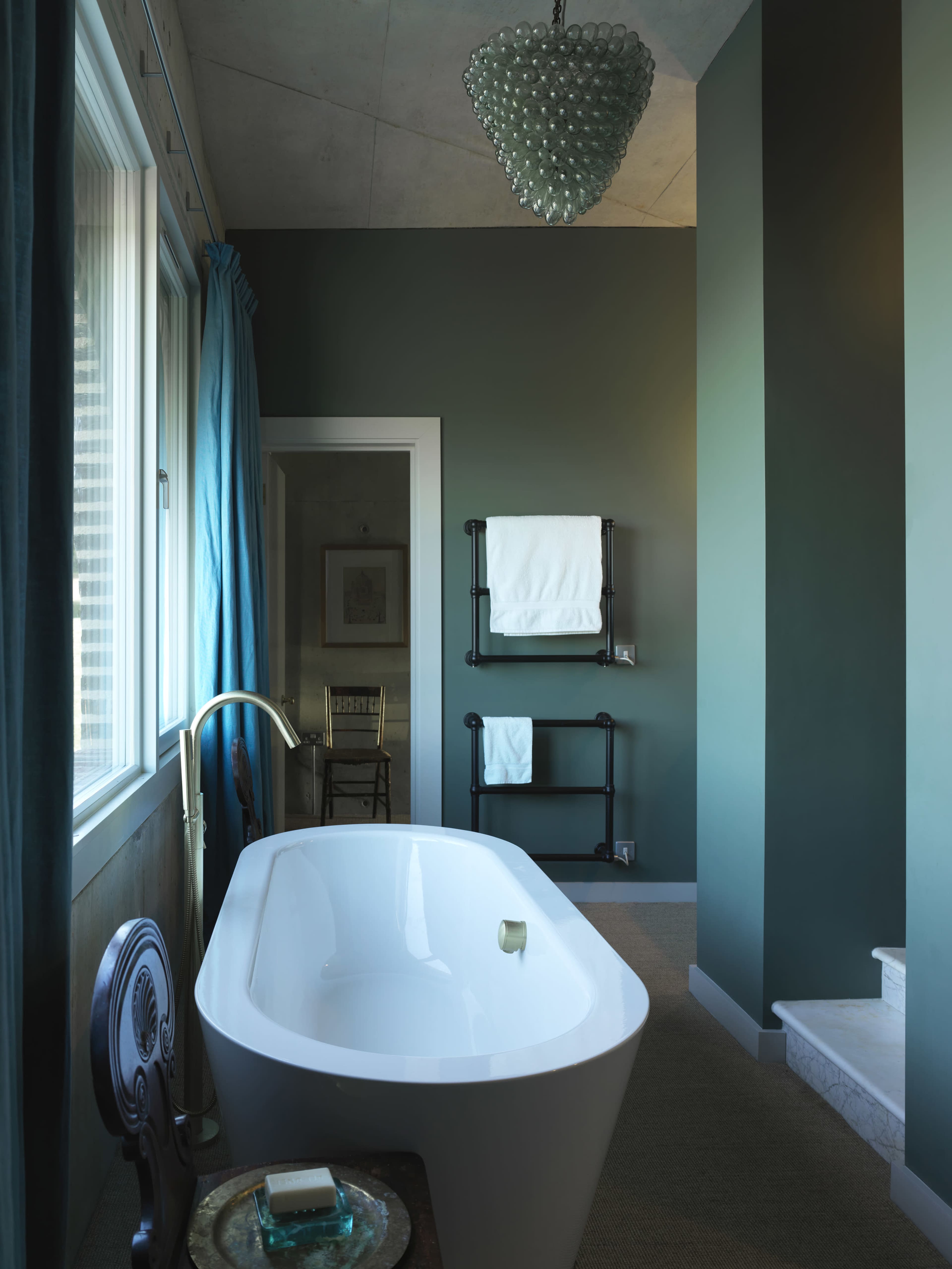A modern bathroom features a freestanding white bathtub, a blue curtain, and a green accent wall, with a chandelier overhead and towels neatly displayed on a black rack.