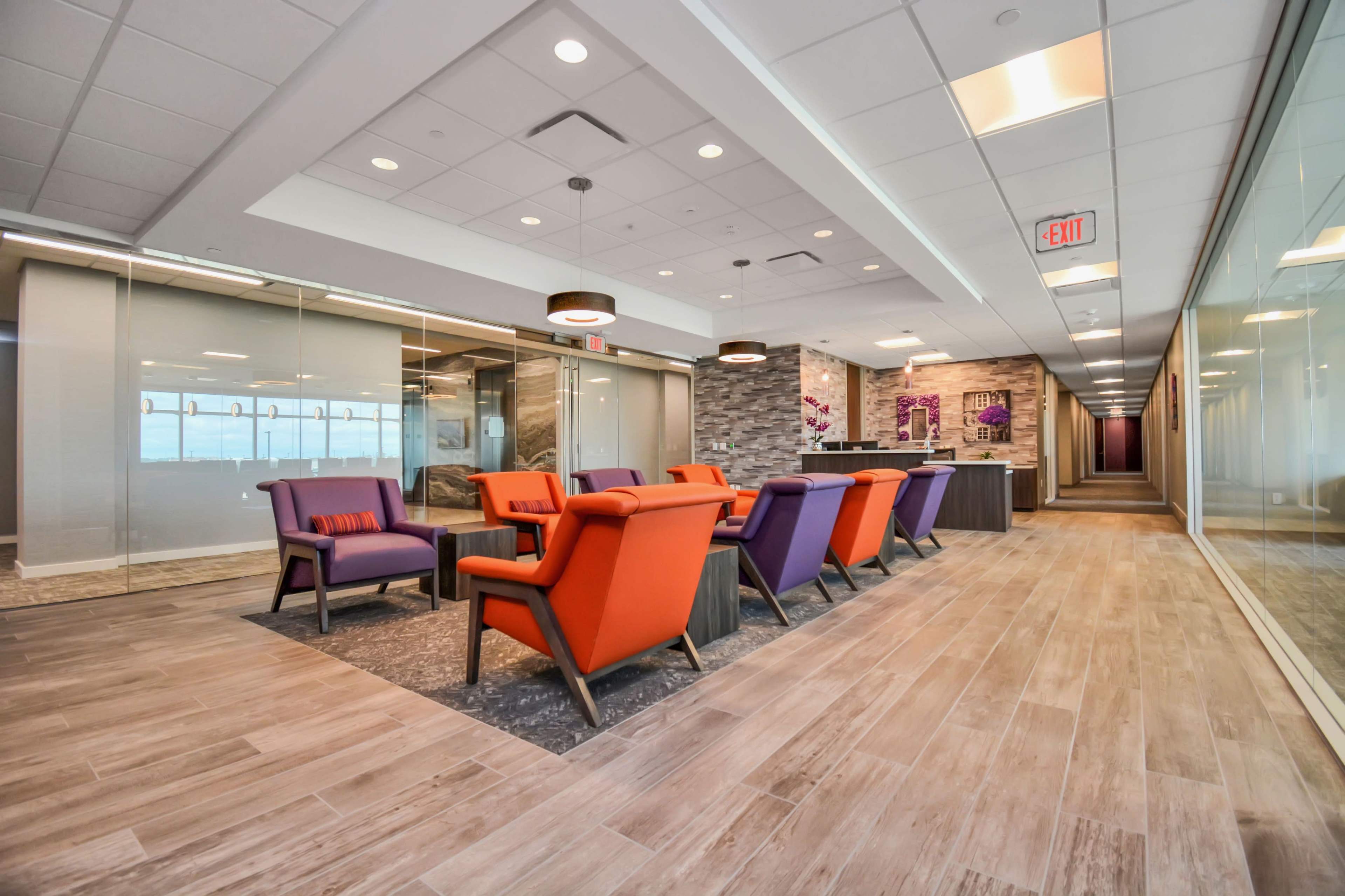 The image shows a modern office corridor featuring colorful lounge chairs arranged around a coffee table, with glass partitions along the walls.