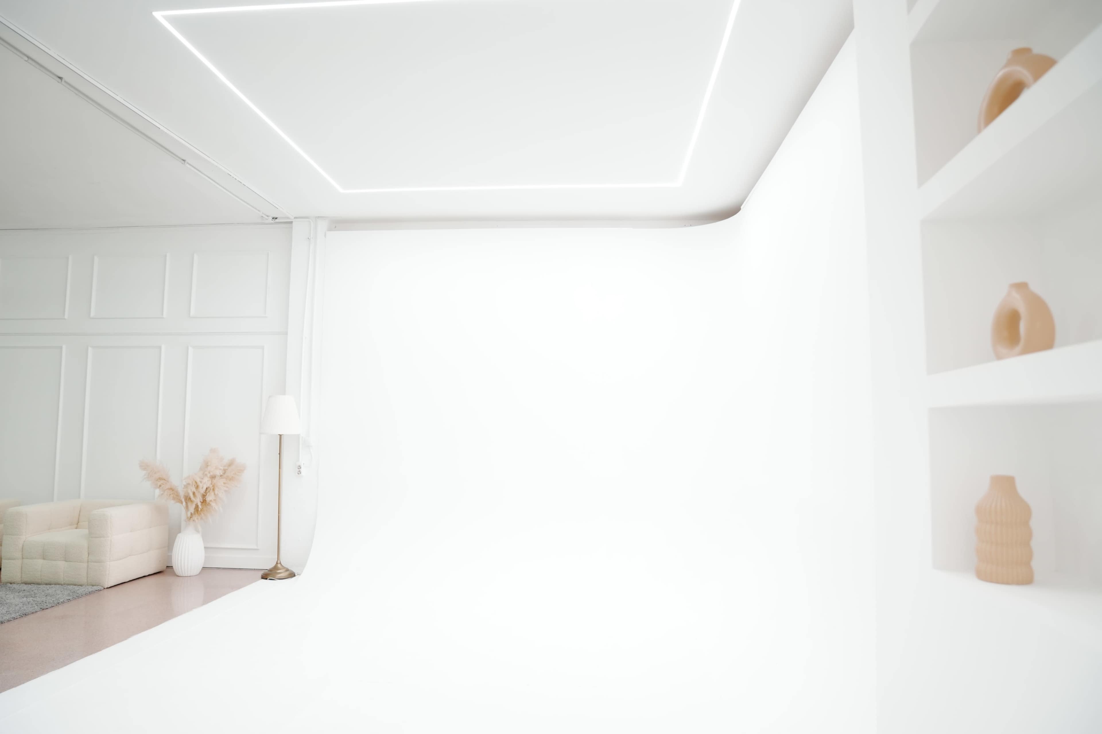 The image shows a minimalist, bright studio space with white walls, a soft beige sofa, a floor lamp, and decorative vases on a shelf.