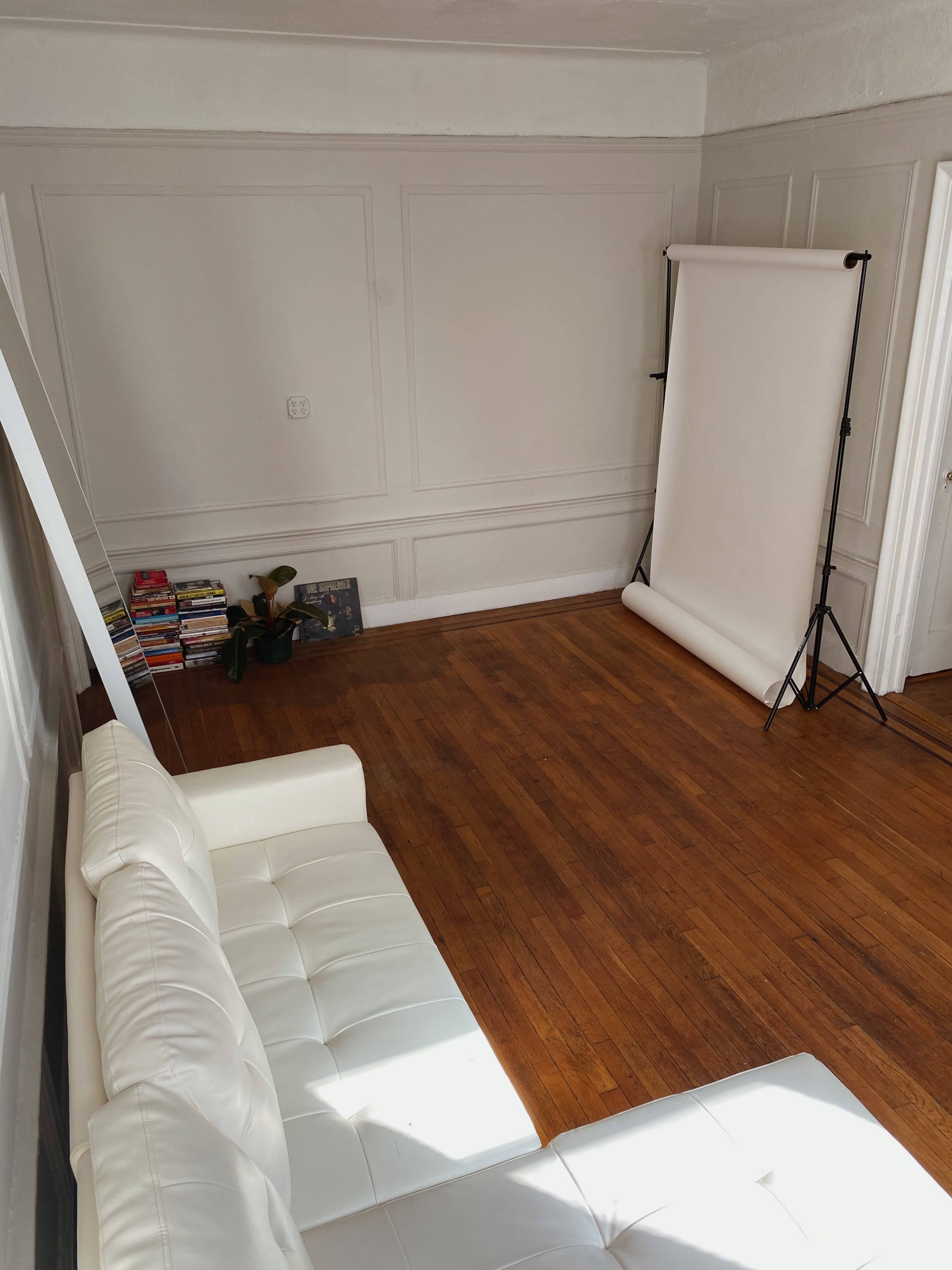 The image shows a minimalist room with a white sofa, a backdrop setup on a stand, and a shelf of magazines against the wall.