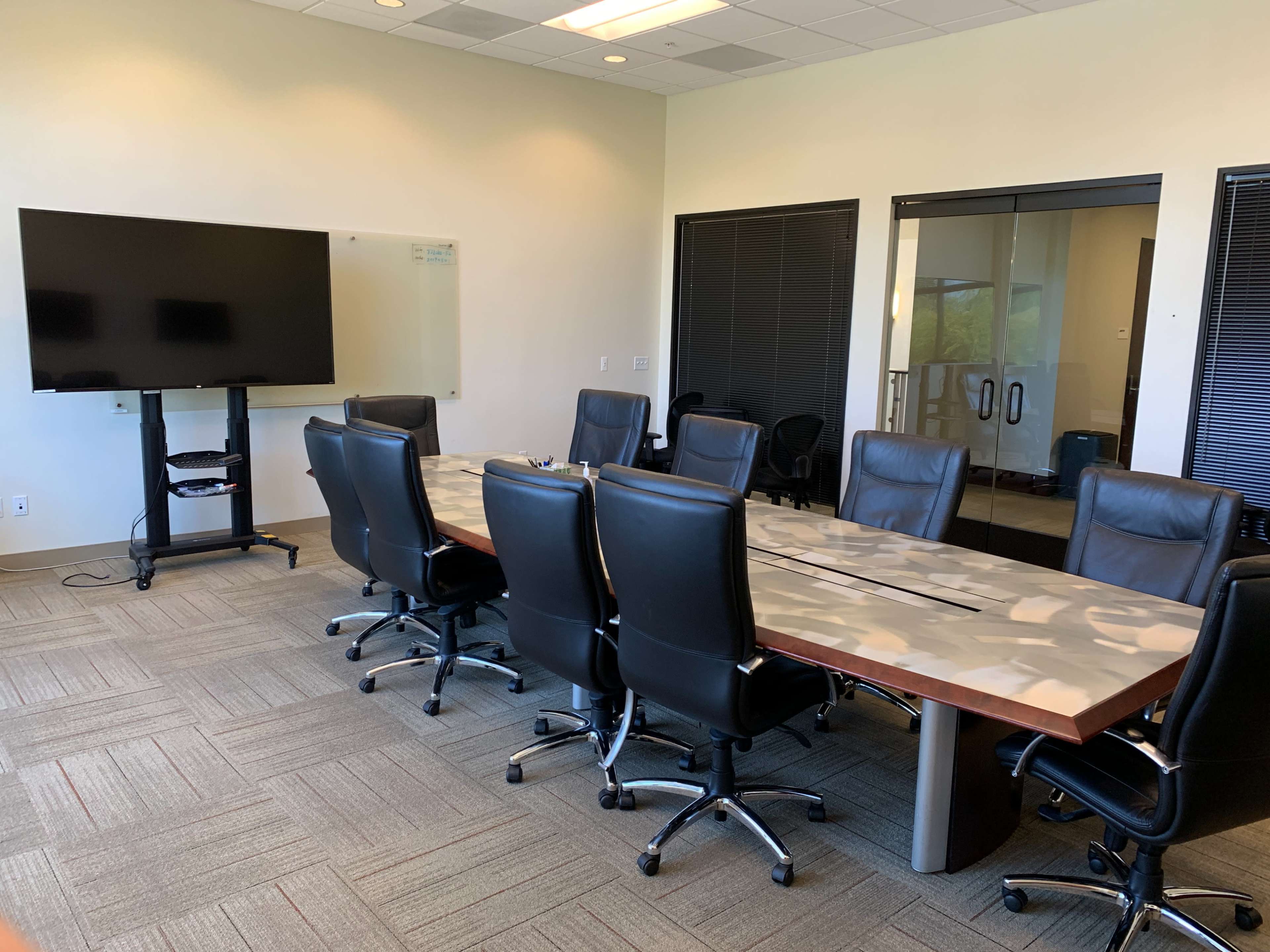 A conference room features a long table surrounded by leather chairs, a large television on a stand, and glass doors leading to another area.