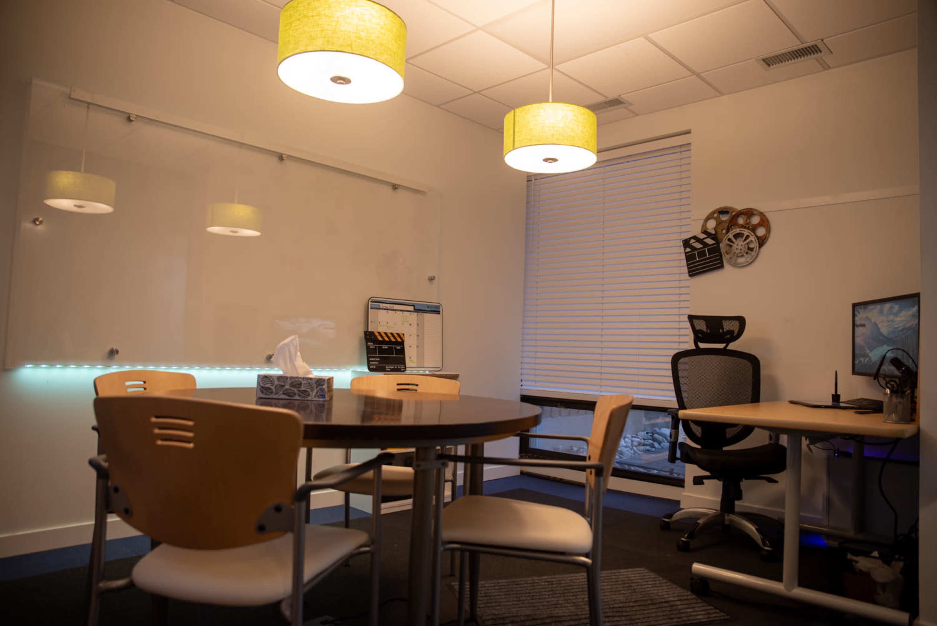 A modern office space features a round table with four chairs, a wall-mounted whiteboard, a calendar, and a desk equipped with a computer and an ergonomic chair.