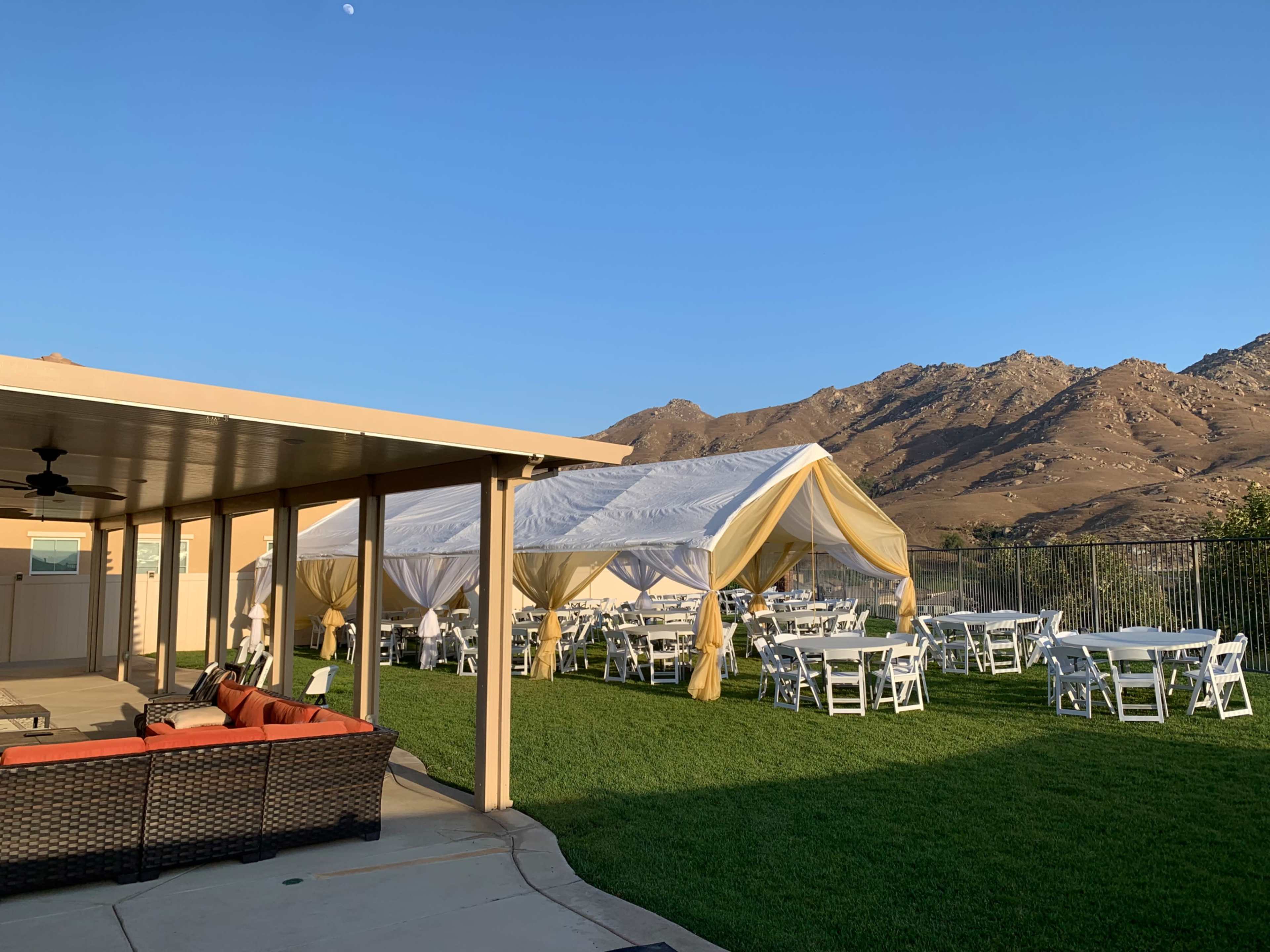 A white tent with yellow accents is set up in a grassy area next to a building, with tables and chairs arranged underneath, surrounded by mountains under a clear blue sky.