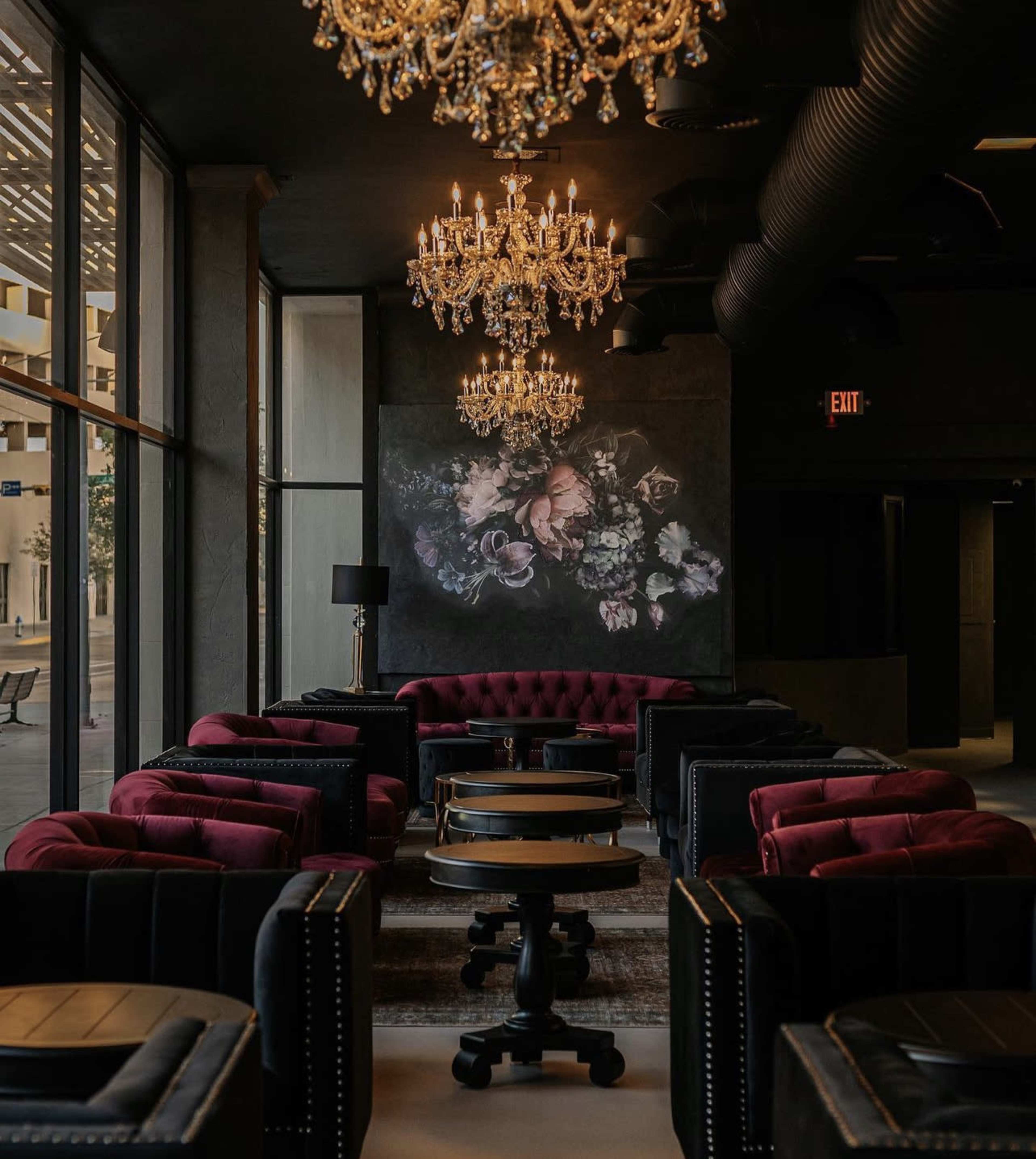 A stylish lounge area featuring plush red and black seating, two ornate chandeliers, and a large floral artwork on the wall.
