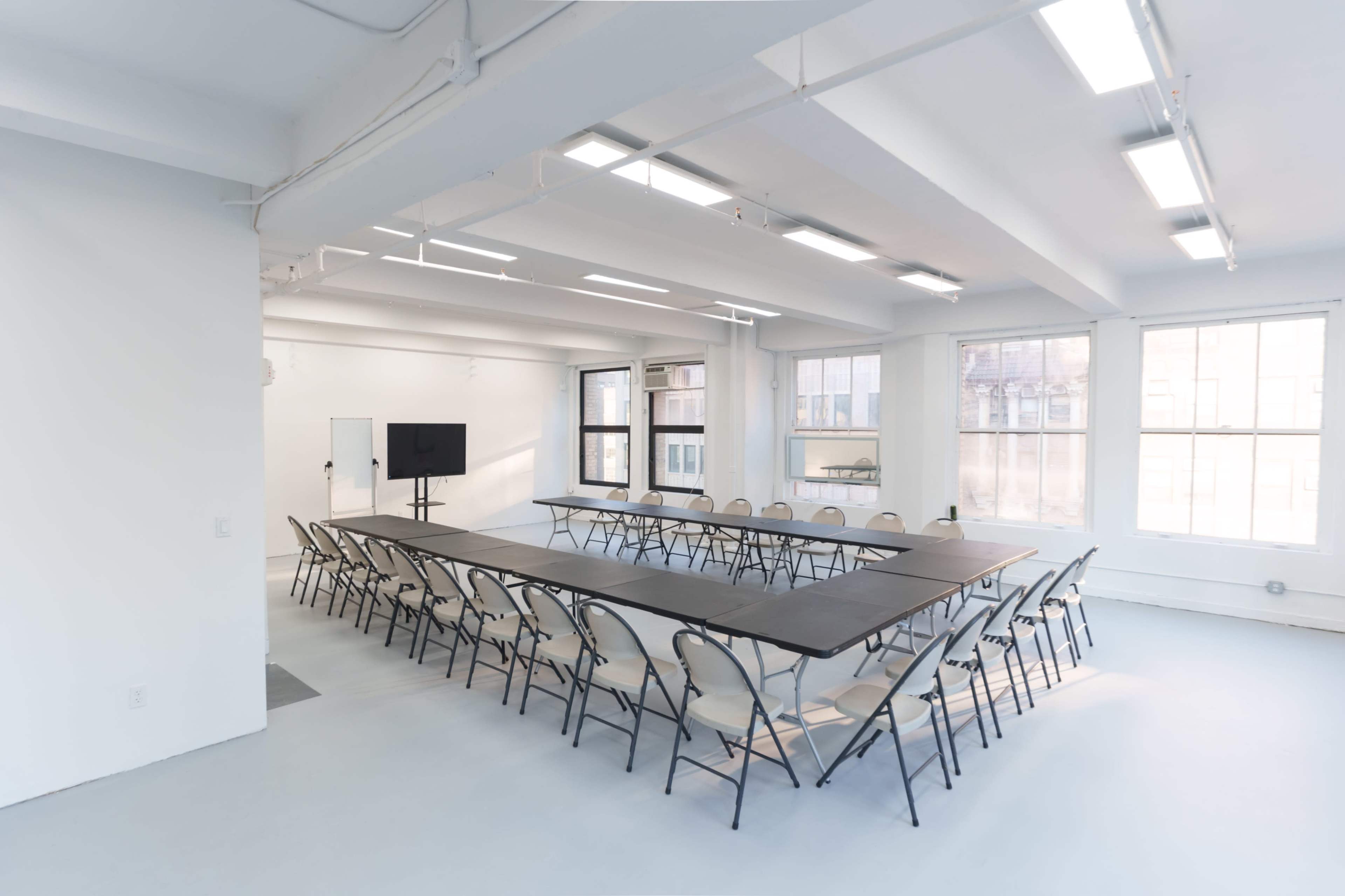 The image shows a modern conference room with a large rectangular table surrounded by folding chairs, bright lighting, and large windows.