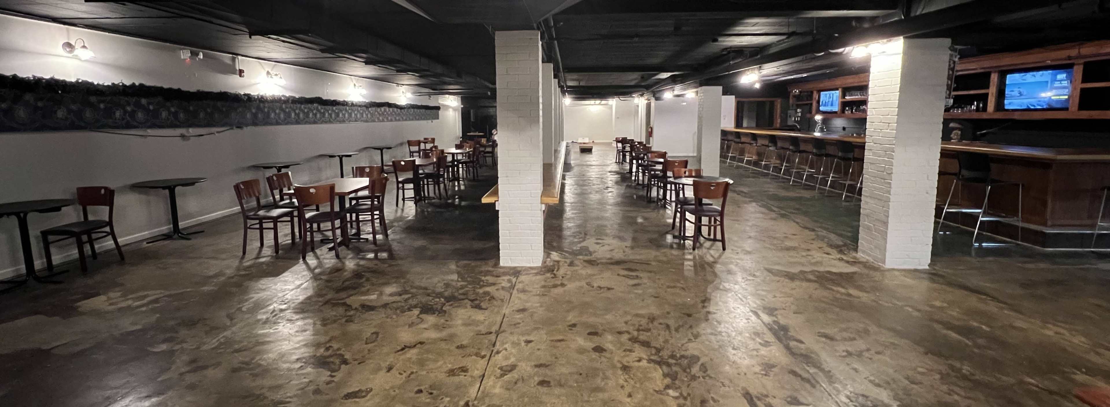 A spacious, dimly lit bar area with metal tables and wooden chairs, featuring a long counter in the background.