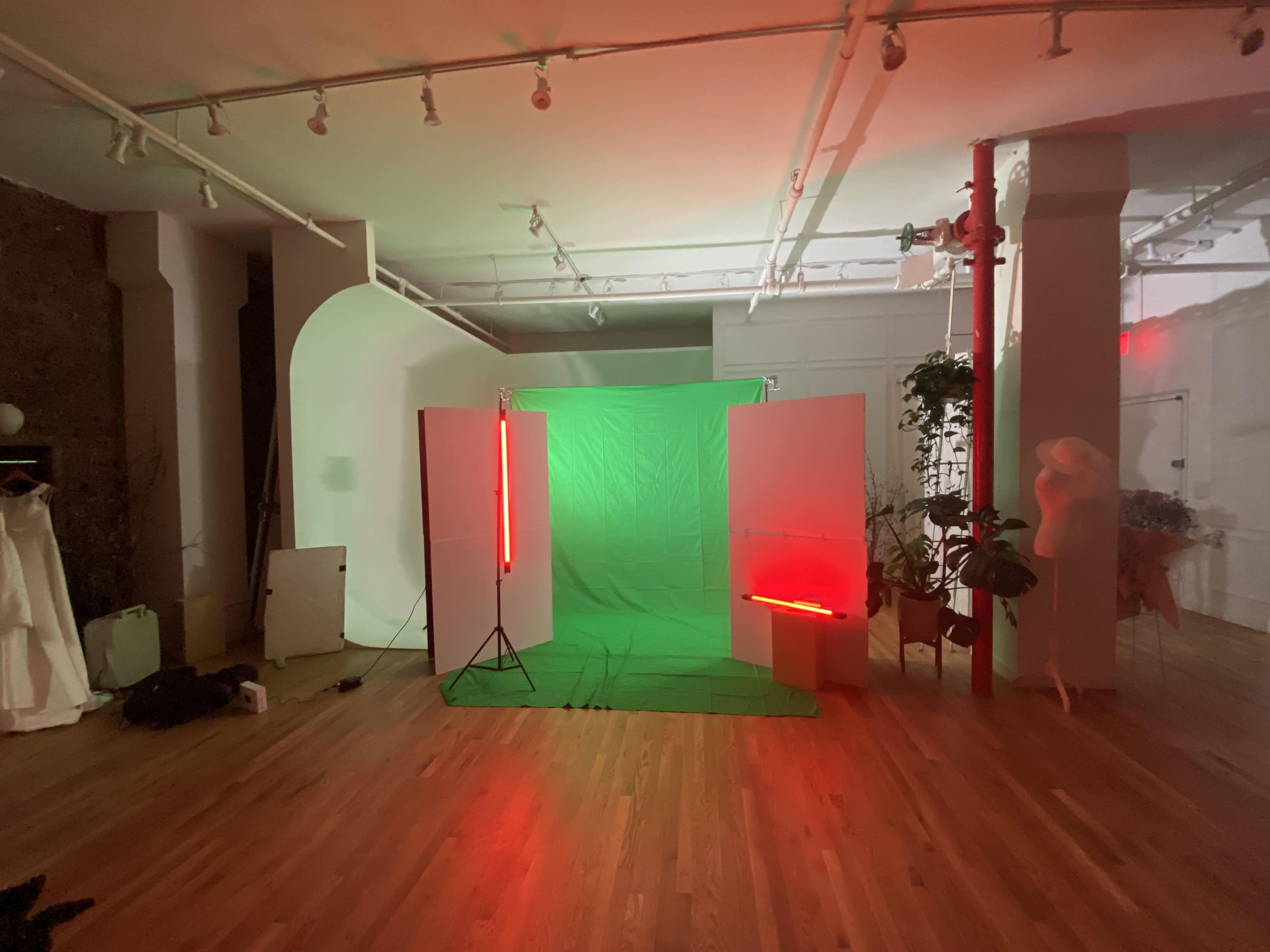 The image shows a studio space with a green backdrop, illuminated by red neon lights, surrounded by white partitions and plants.