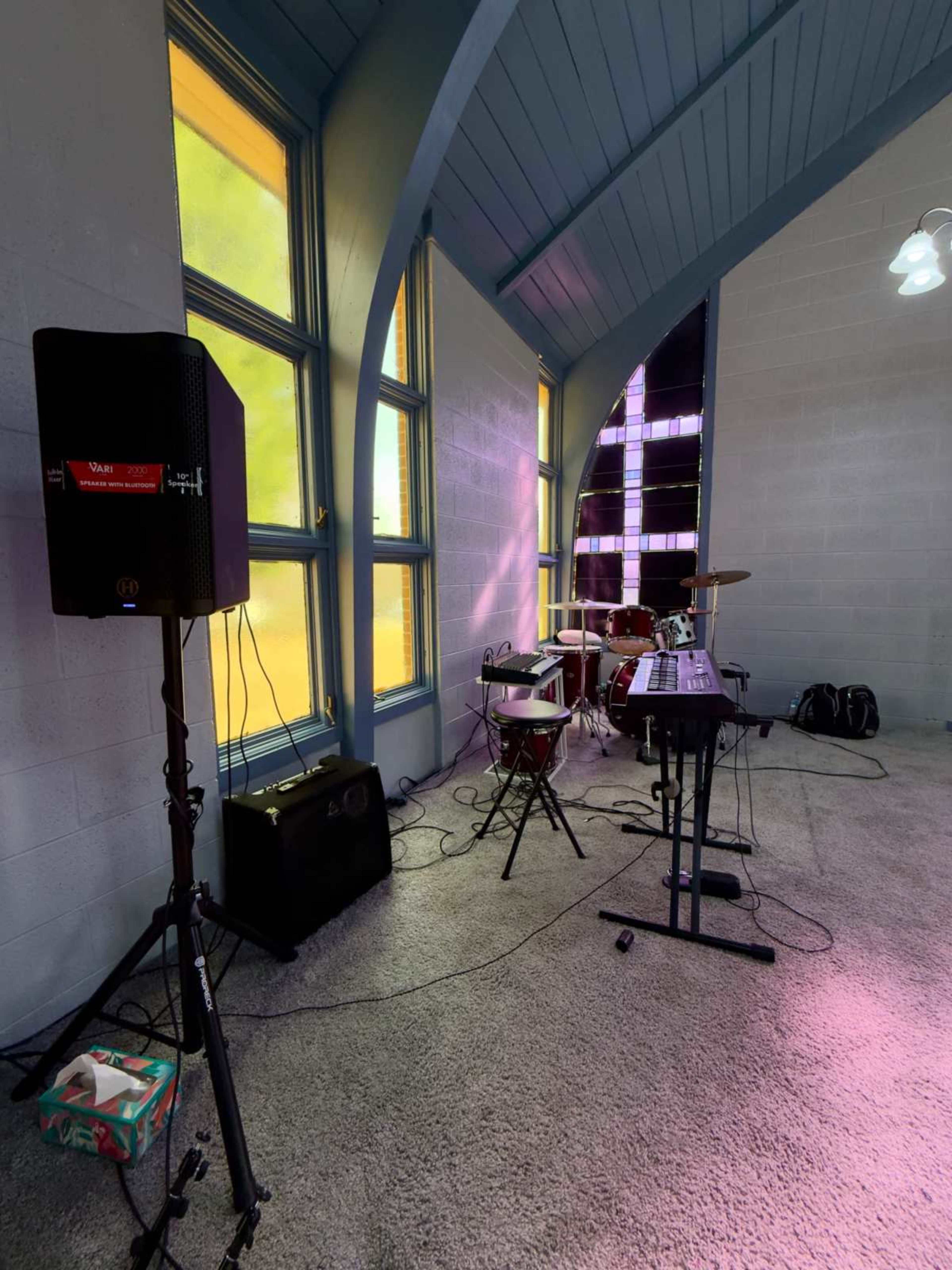 The image shows a room with colorful windows, housing a drum set, keyboard, and amplifiers on stands.