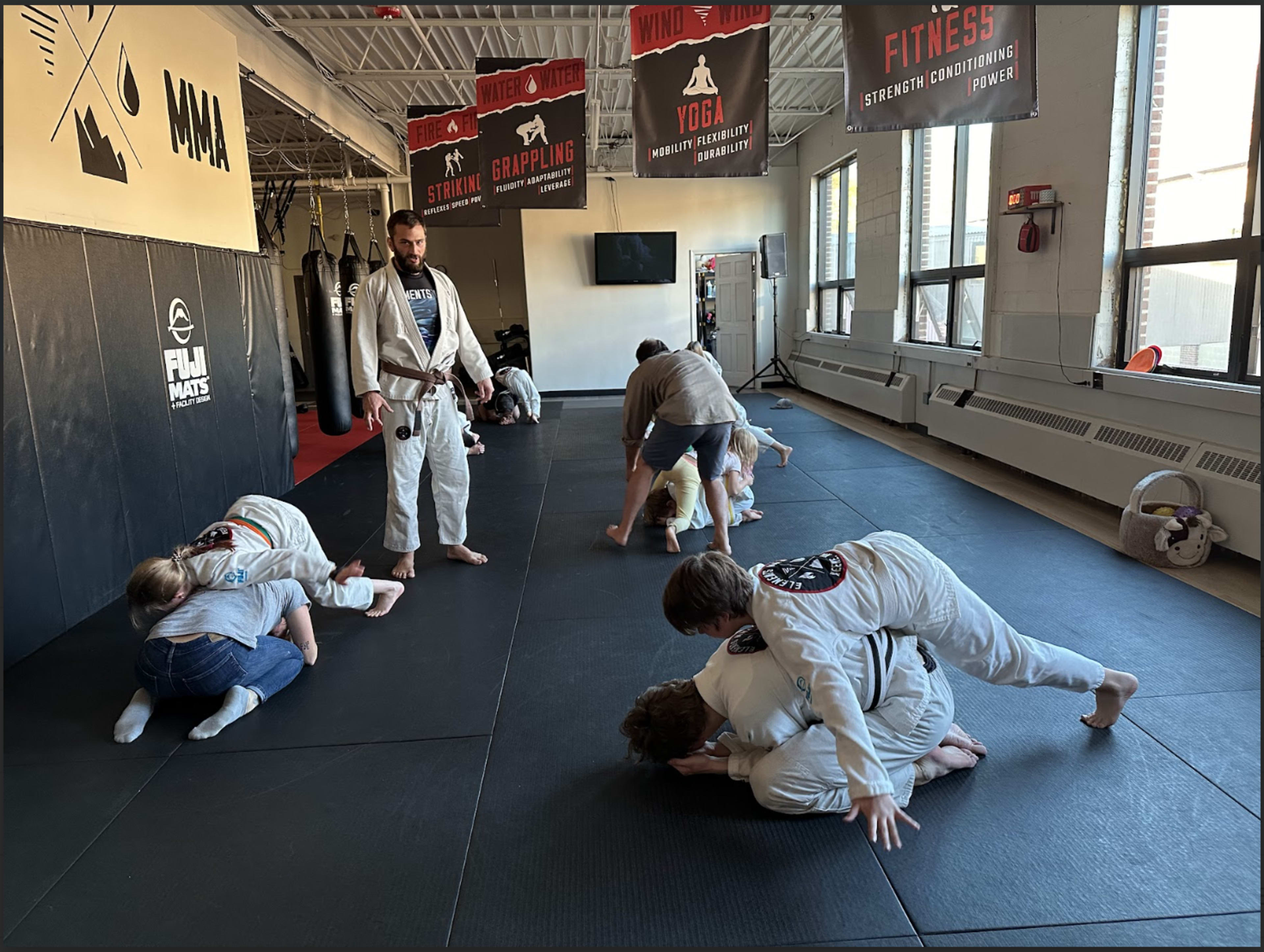 Several individuals wearing gi uniforms are practicing jiu-jitsu techniques on a black mat inside a gym, while an instructor oversees their training.