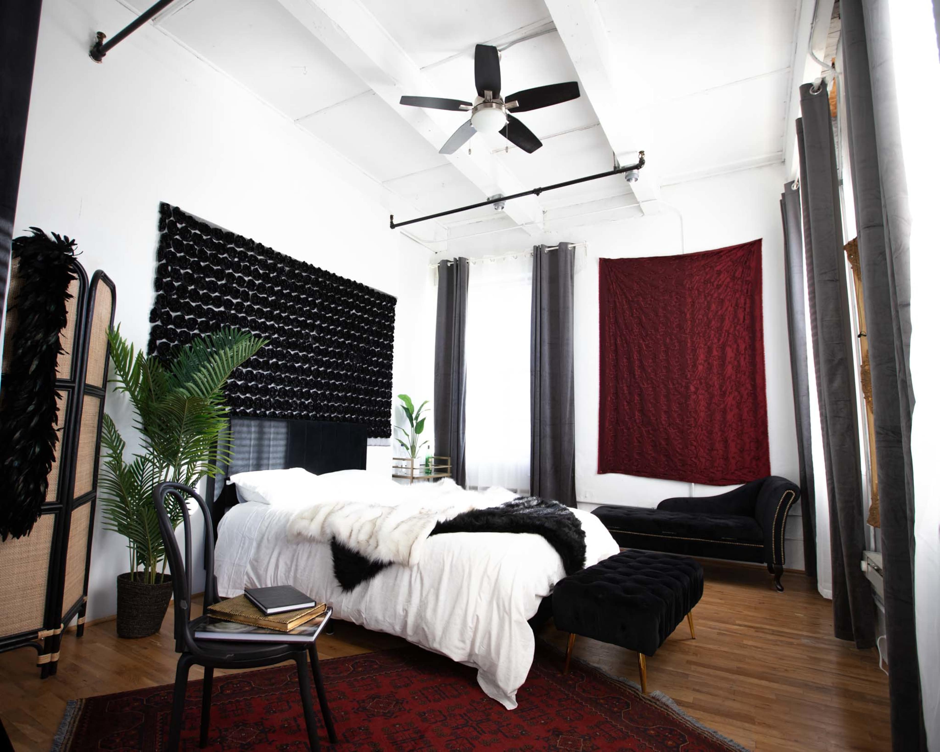 The room features a bed with a black headboard, surrounded by potted plants and a vintage chaise lounge, all set against white walls and hardwood flooring.