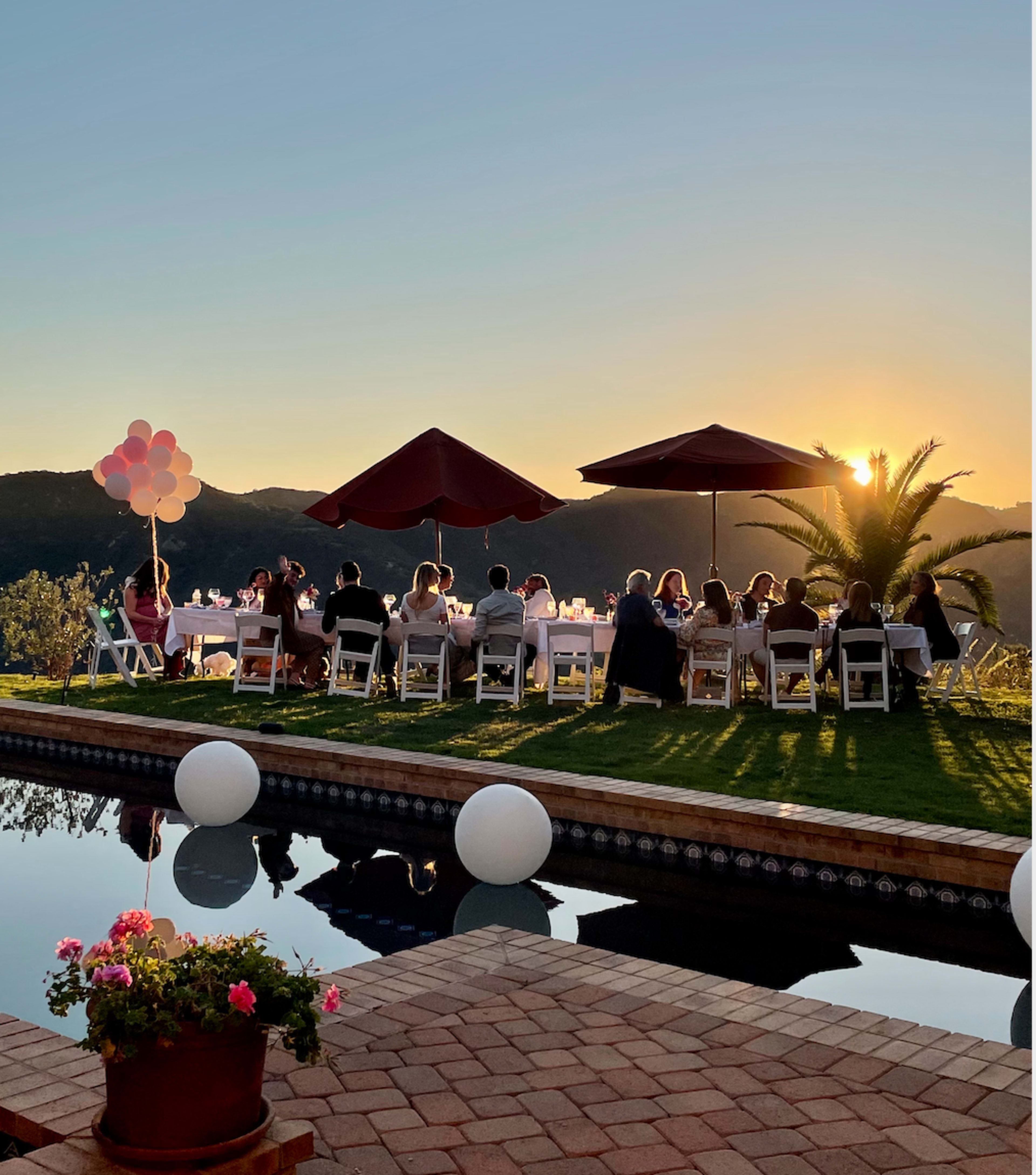 A group of people sit around a long table with umbrellas at a sunset gathering beside a pool, with balloons and a scenic landscape in the background.