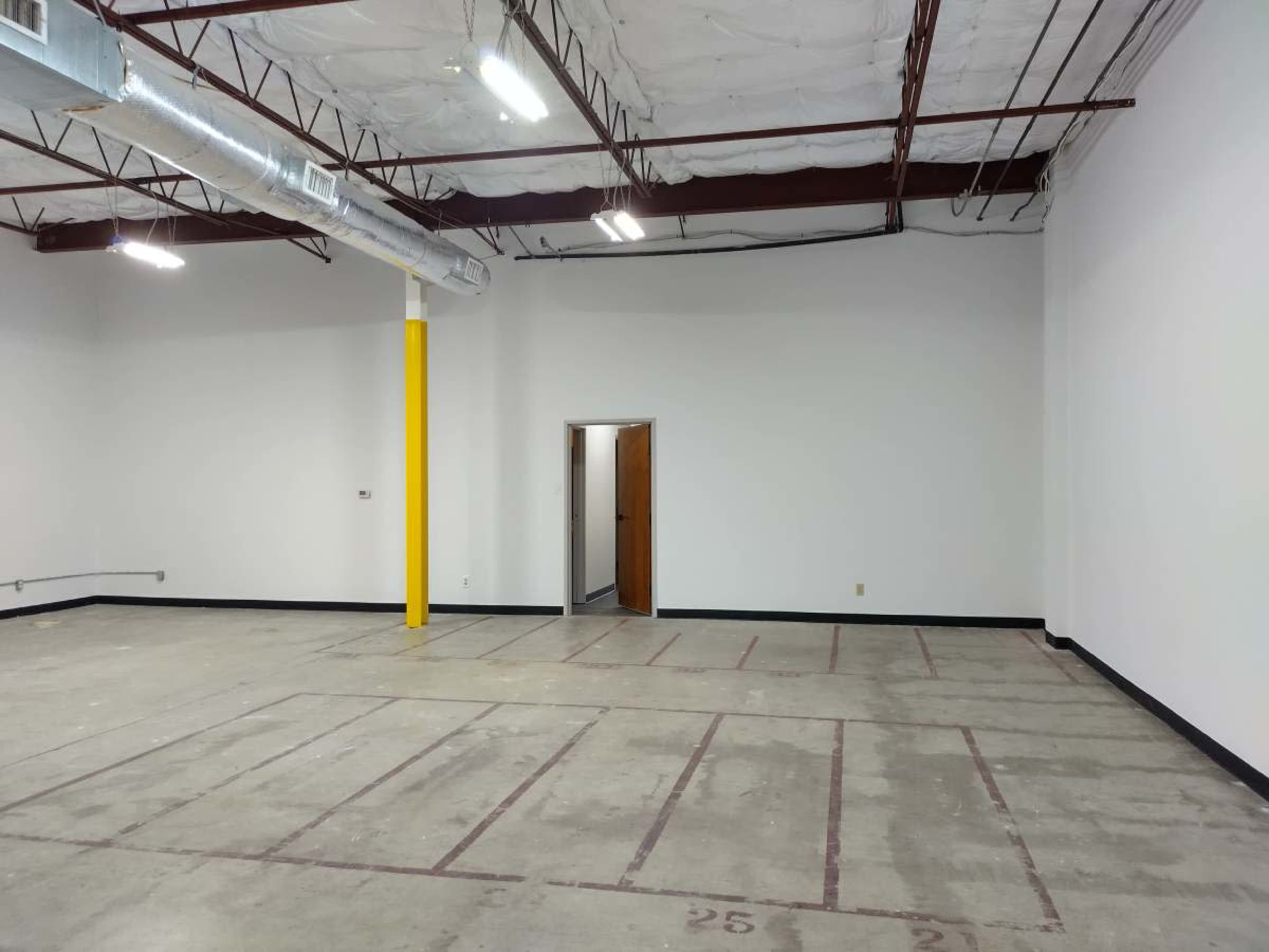 The image shows an empty room with concrete flooring, white walls, exposed ceiling beams, and a door leading to another space.