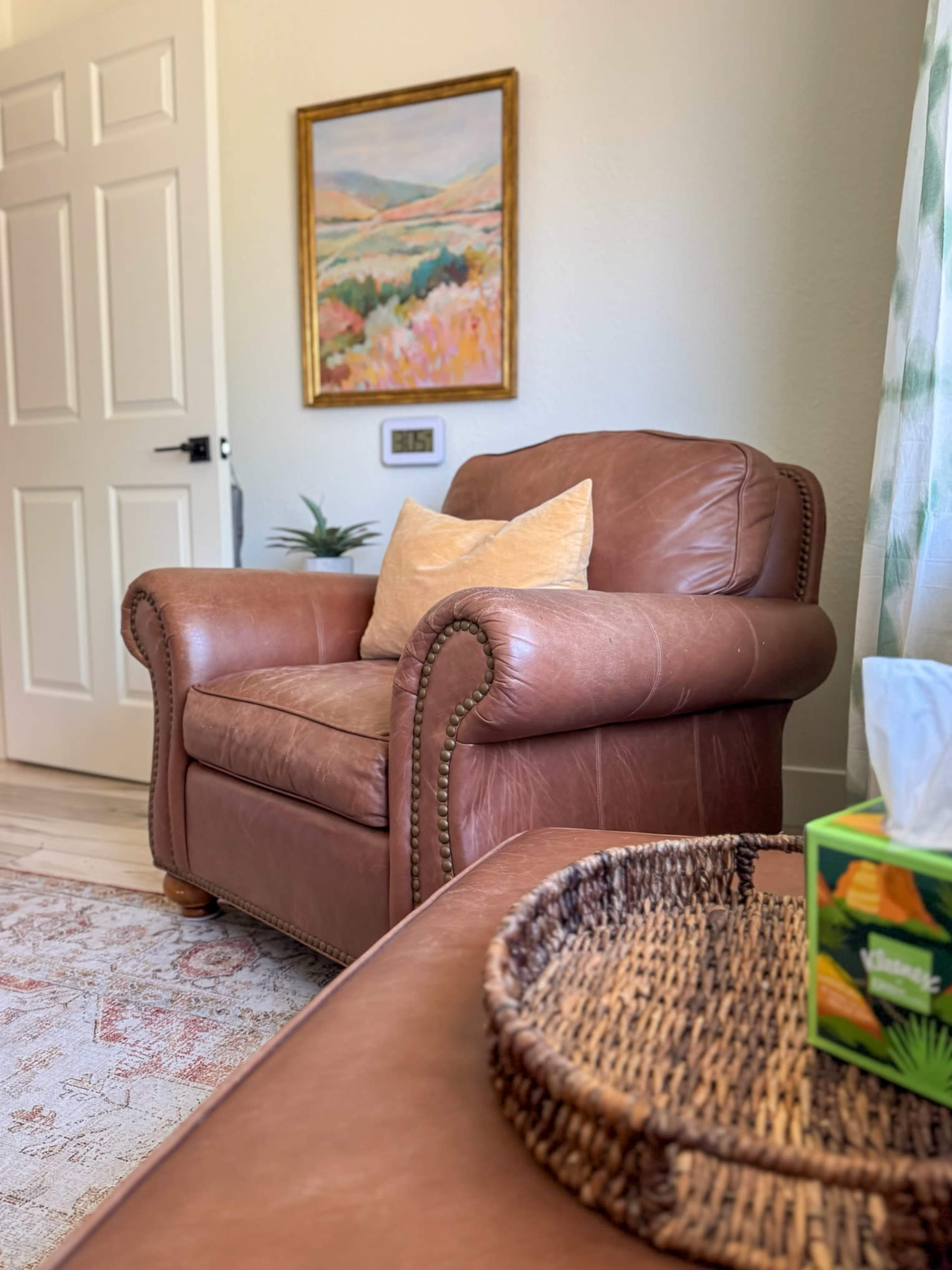 A brown leather armchair is positioned near a door and a wall with a painting, while a woven tray holds a box of tissues and is placed on a nearby surface.