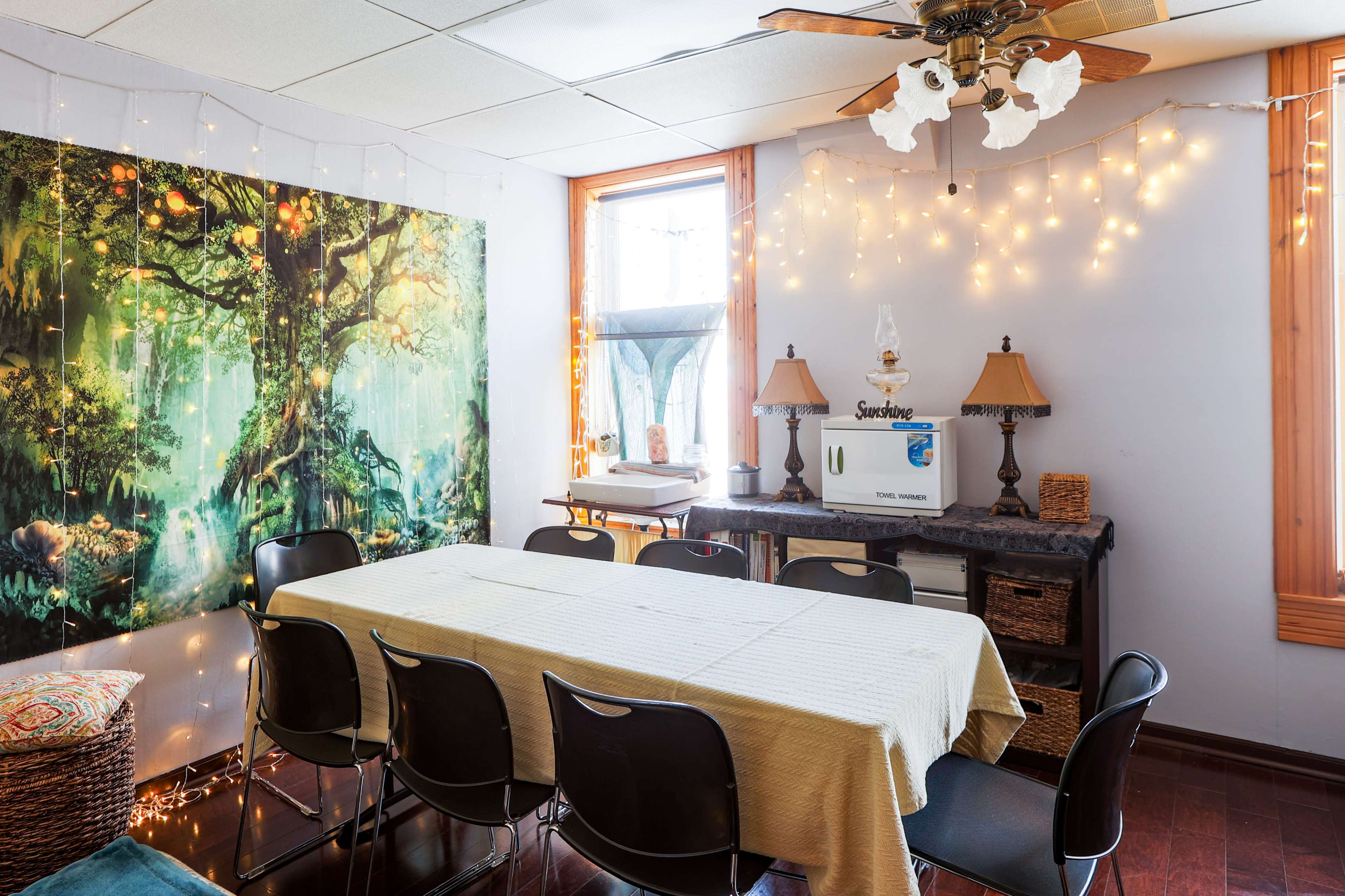 The image shows a cozy dining area with a long table surrounded by black chairs, illuminated by string lights and adorned with a vibrant wall mural.