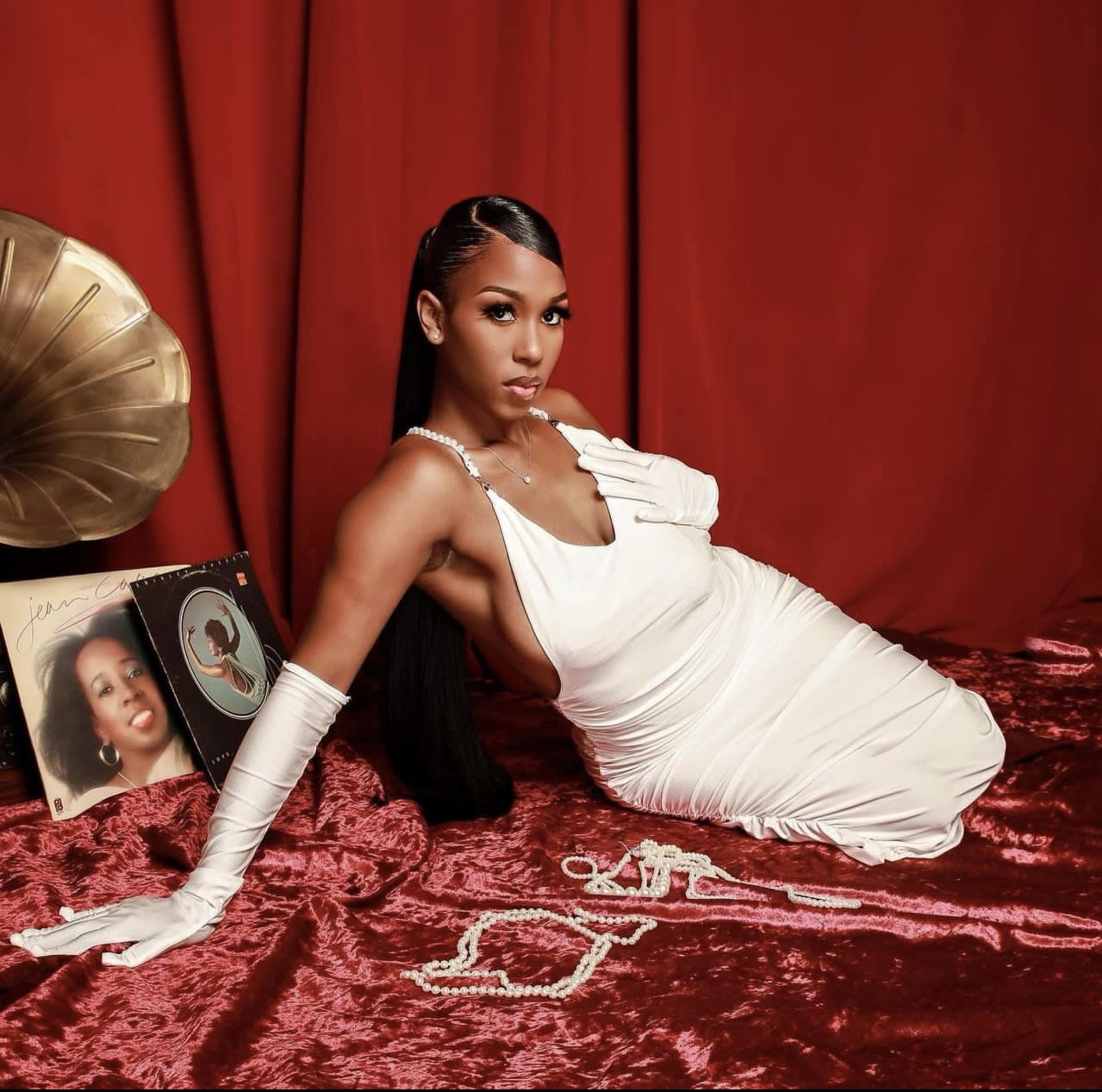 A woman in a white dress poses elegantly on a red surface, surrounded by vintage records and jewelry.