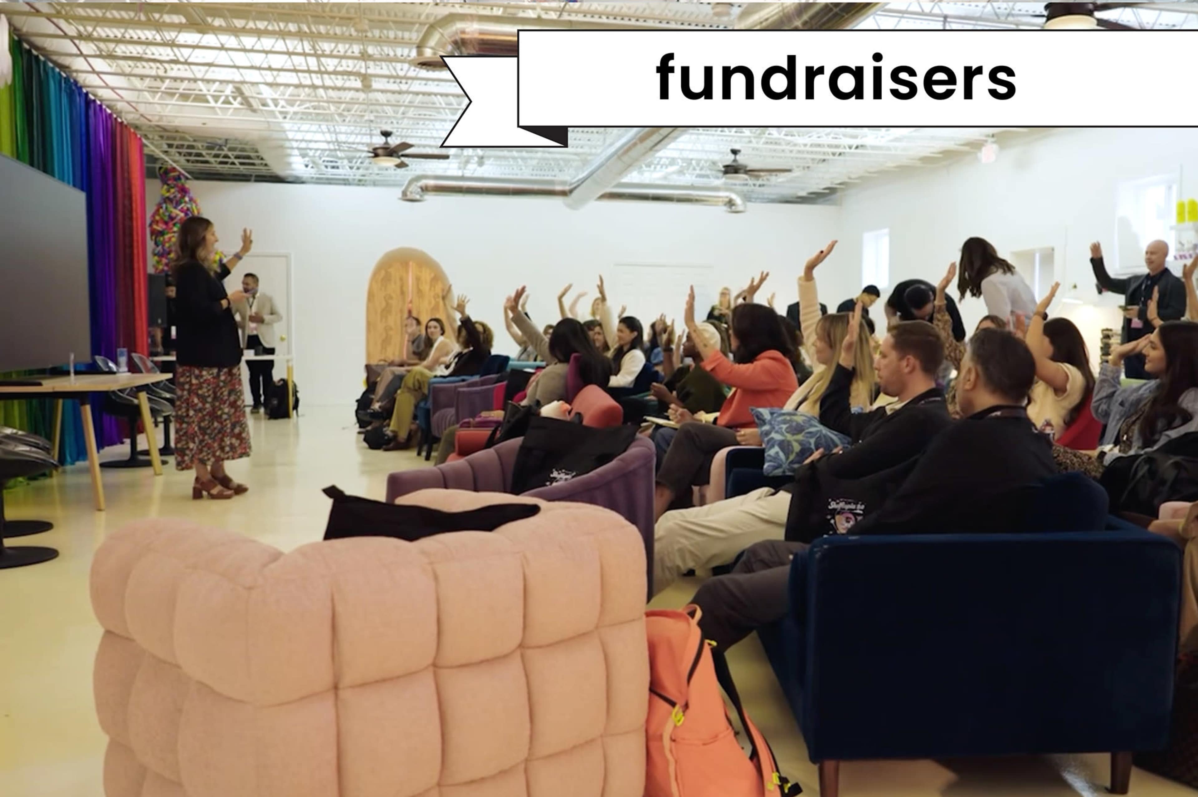 A group of people is seated in a room, with many raising their hands in response to a presenter at the front.