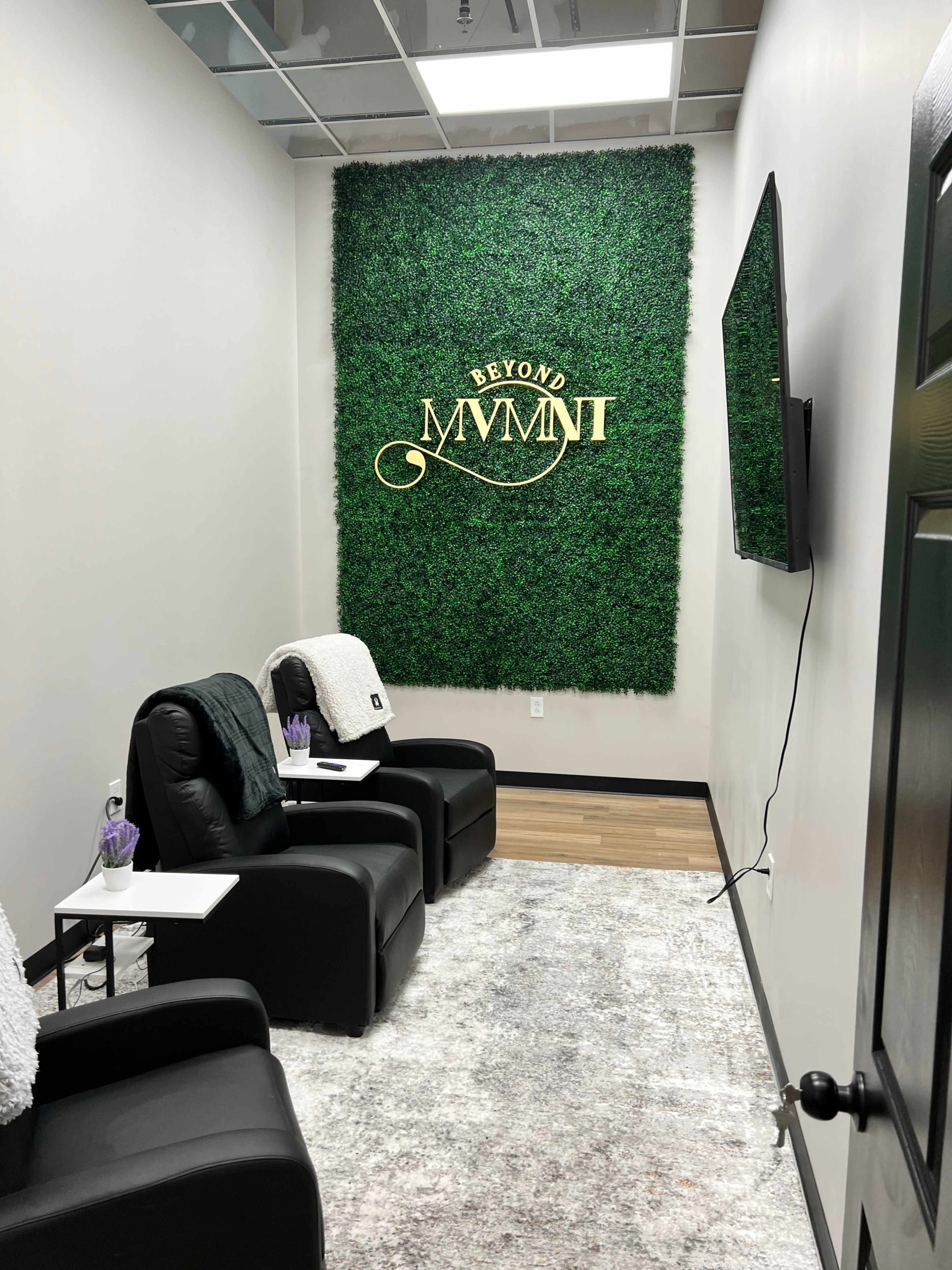 A small room designed for relaxation, featuring two black reclining chairs, a greenery wall with the words "BEYOND MVMT," and a television mounted on the wall.