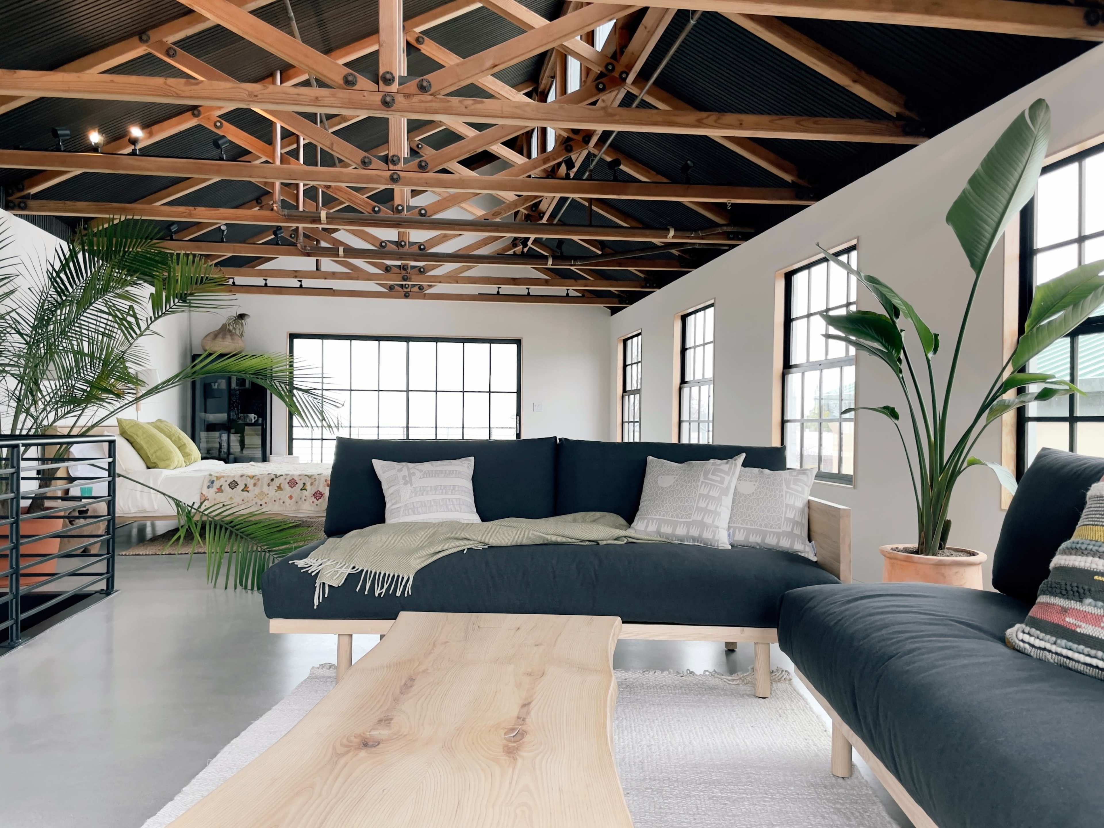 A spacious, industrial-style living room with exposed wooden beams, large windows, and modern furniture, including a dark sectional sofa and a wooden coffee table.