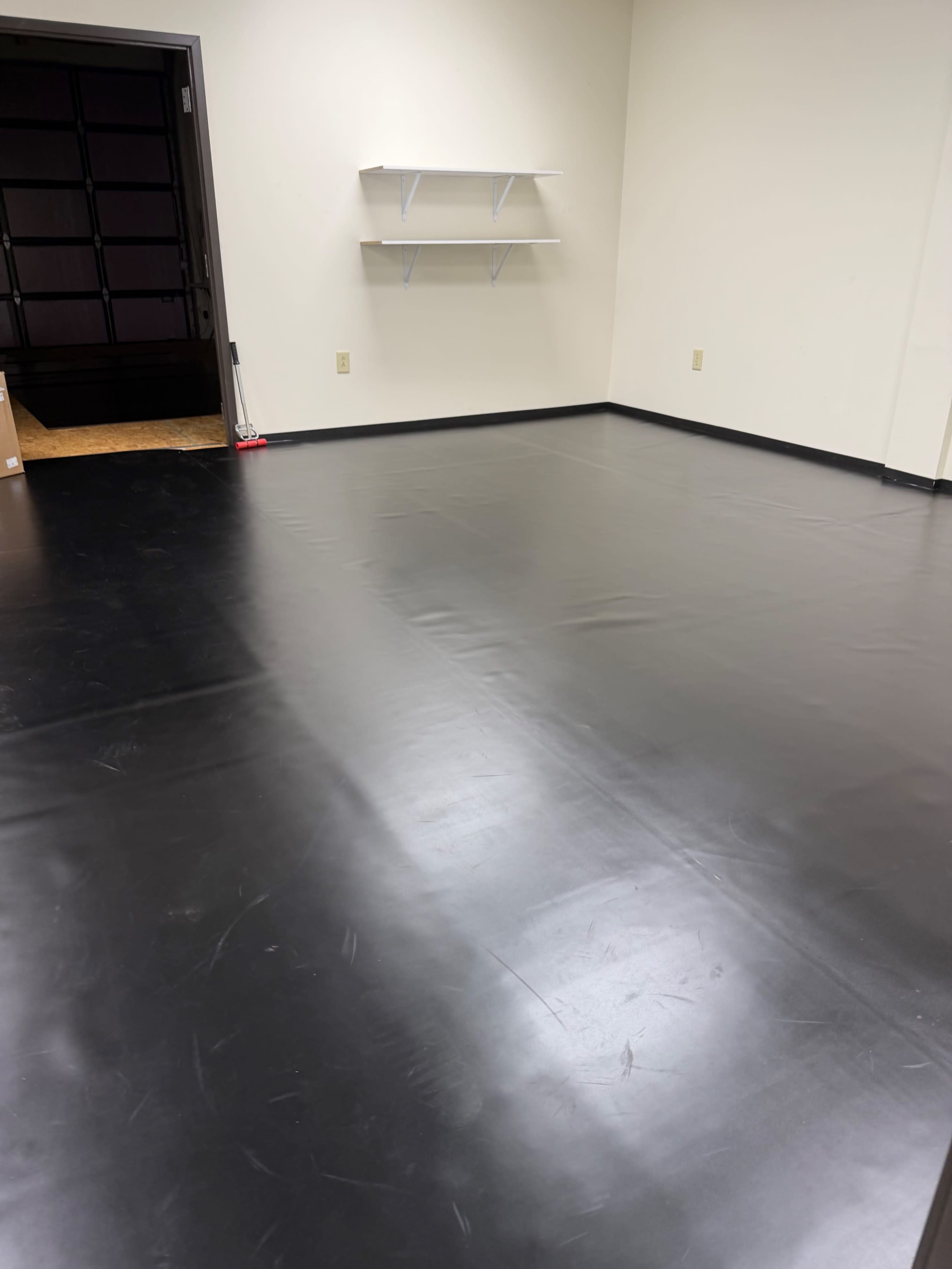 The image shows a sparsely furnished room with a black vinyl floor, featuring a bare wall with two shelves and an entrance door visible in the background.